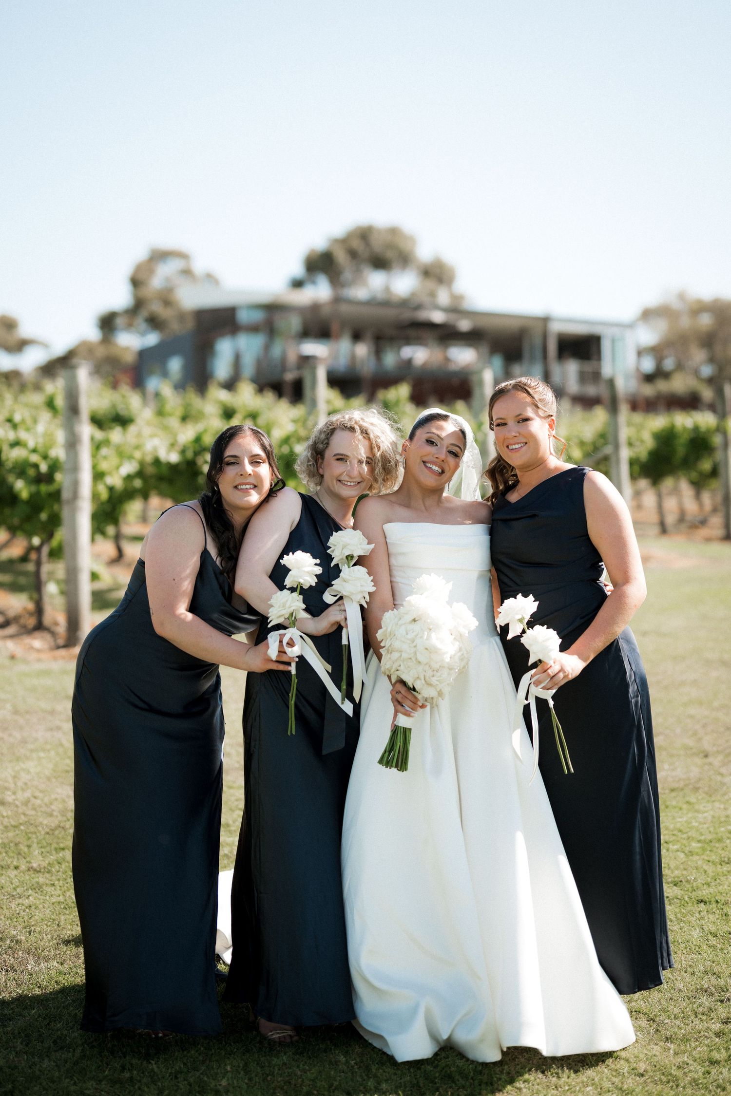 Wedding party in black dresses and white gown pose together in an outdoor vineyard setting.