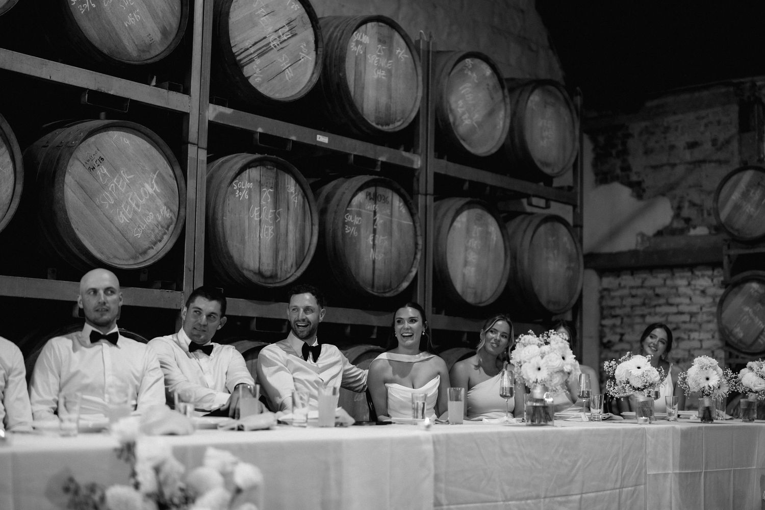 A black and white photograph of a formal dinner gathering with wine barrels stacked along the wall in the background.