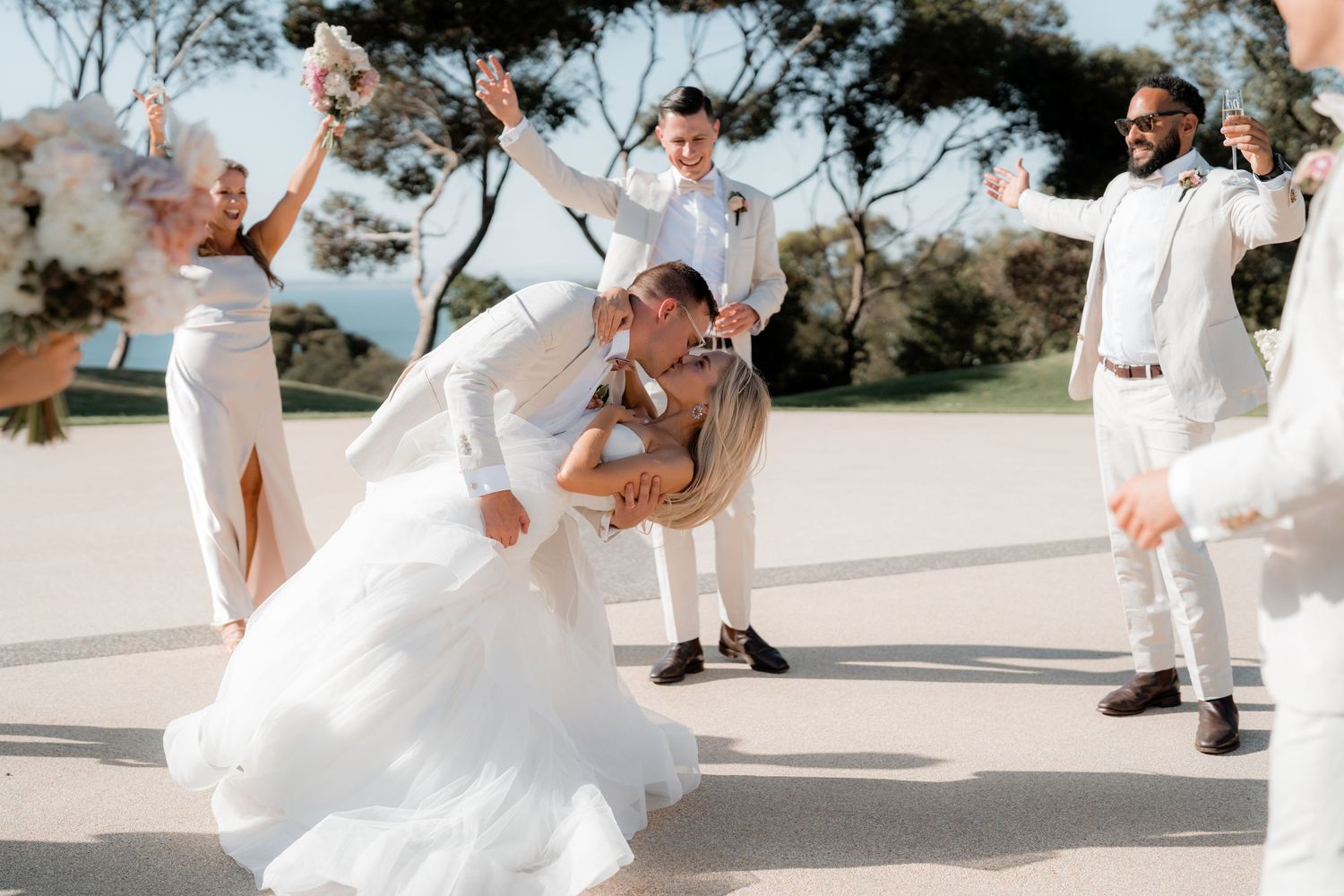 Wedding party in white attire posing outdoors on a sunny day.