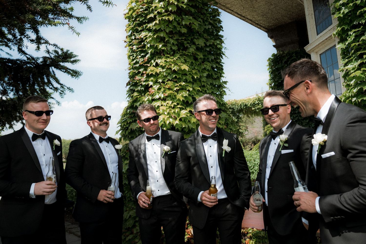 Groomsmen in black tuxedos and sunglasses share drinks and laughs outdoors during a wedding celebration.