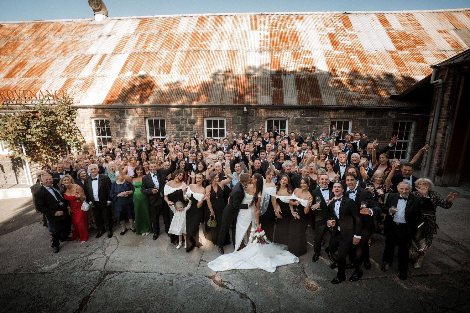 A panoramic wedding photo showing guests gathered outside a rustic brick building with a copper-colored roof.