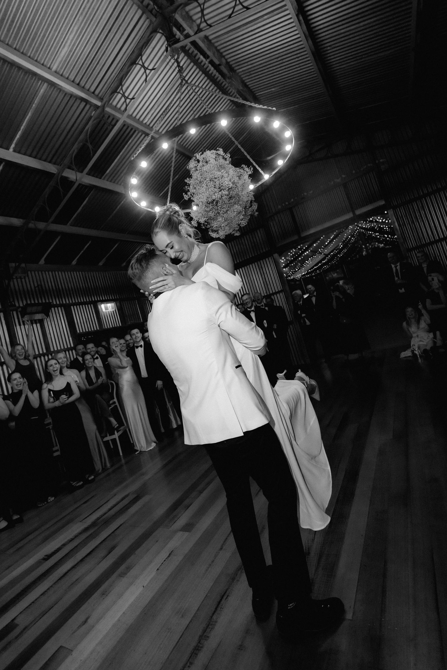 A couple shares a romantic dance moment at a wedding reception in a dimly lit barn venue with string lights overhead.