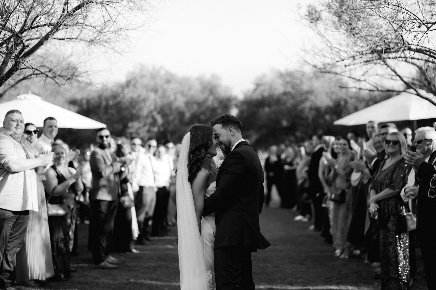 A romantic black and white wedding photo showing a couple surrounded by guests at an outdoor ceremony.