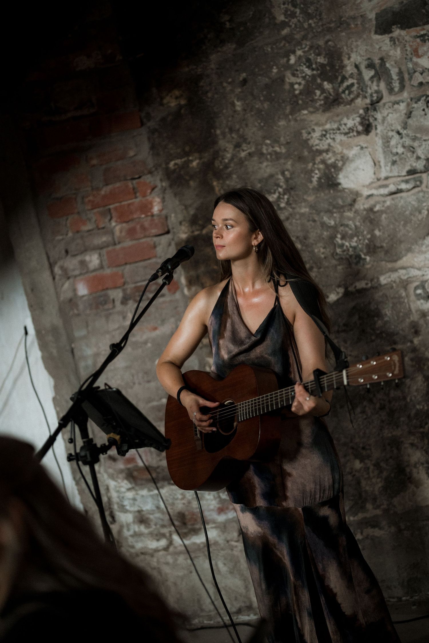 A musician performs with an acoustic guitar against a textured stone wall in moody lighting.