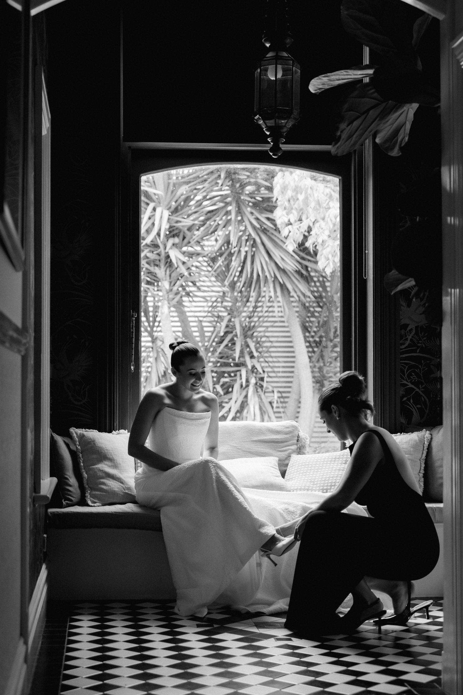Bride in white gown sits as bridesmaid adjusts her shoes in elegant black and white wedding preparation photo.