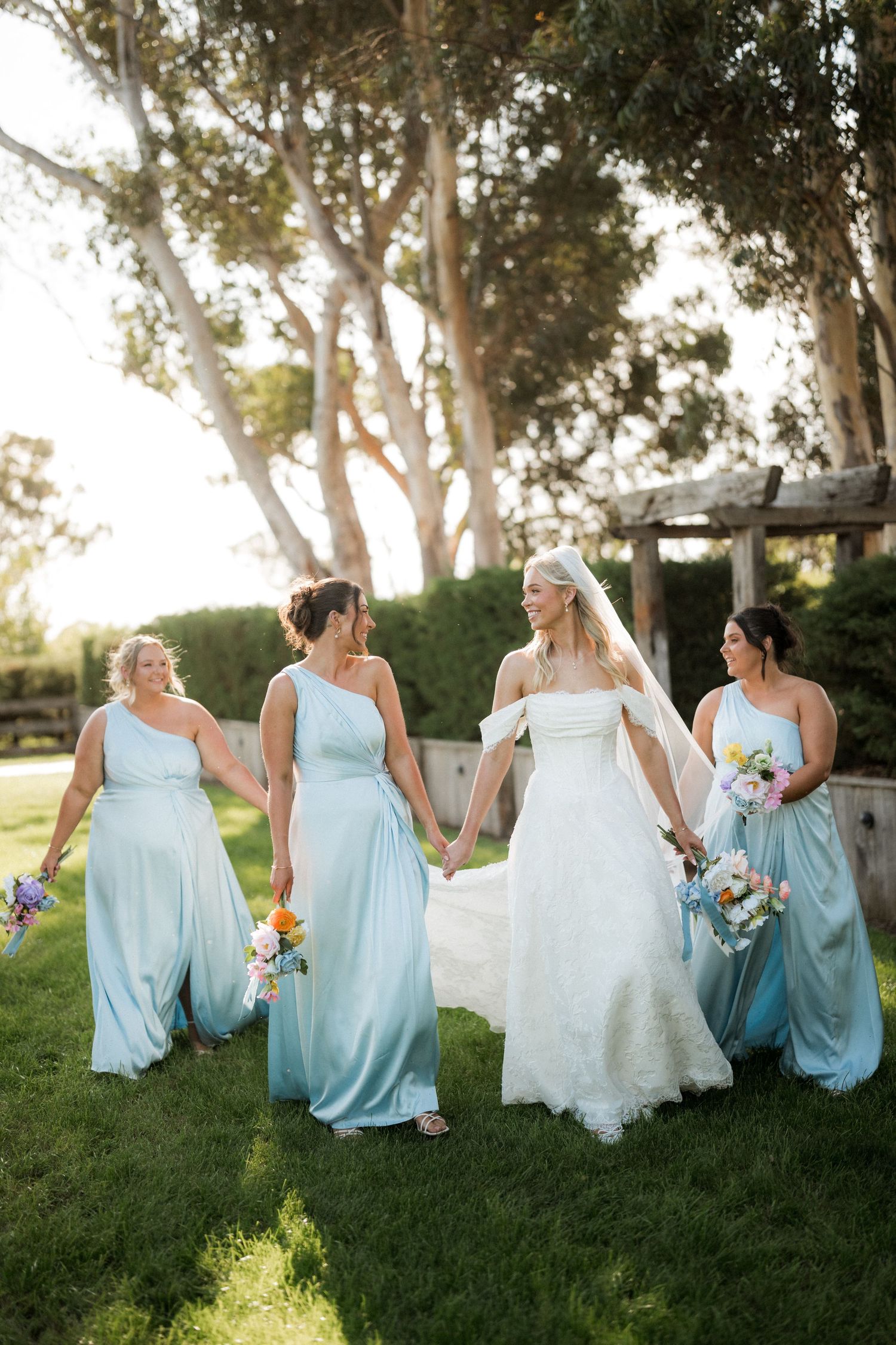 Bride in white gown walks hand-in-hand with three bridesmaids in light blue dresses across a sunlit garden.