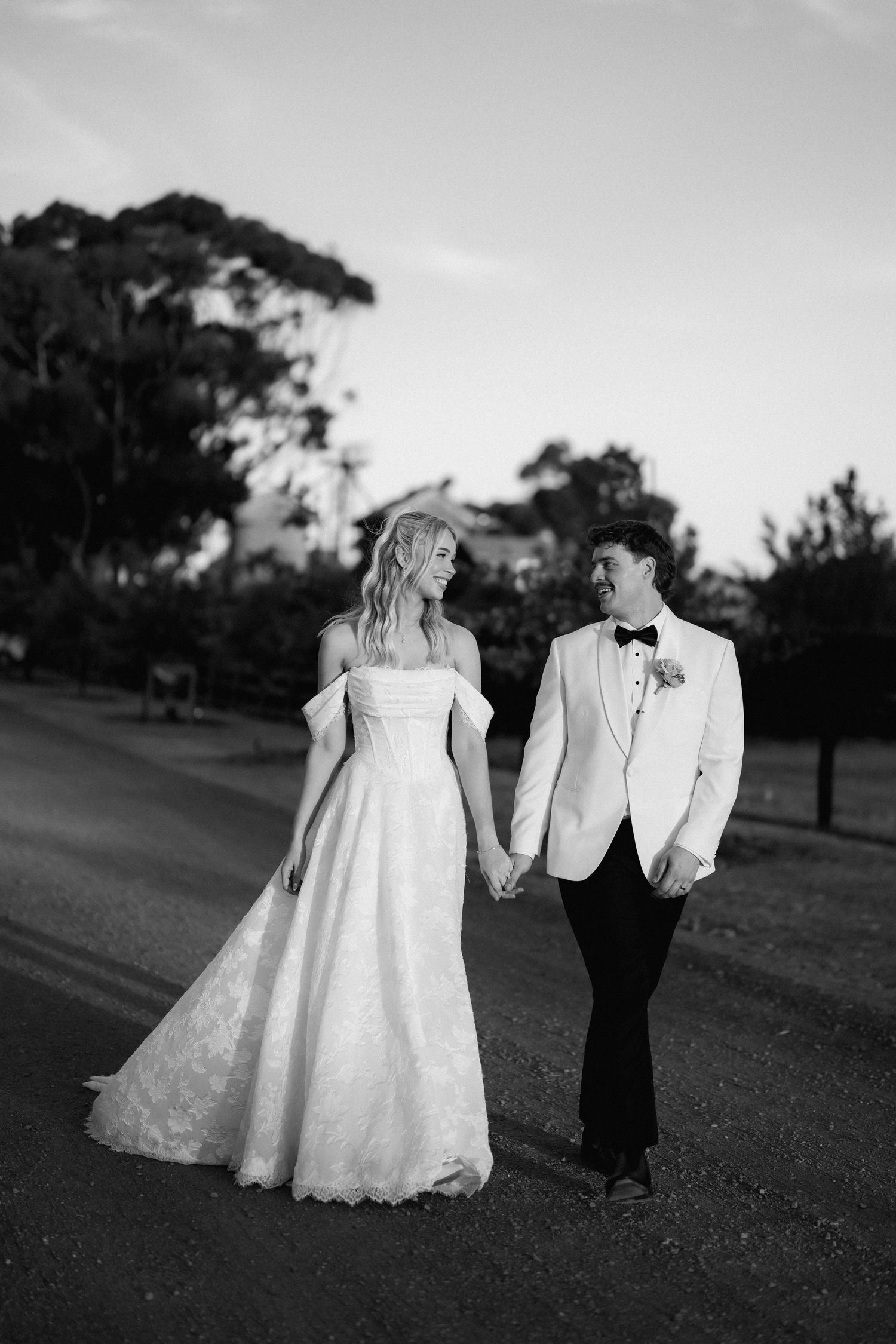 Black and white photo of bride and groom holding hands walking along tree-lined road at dusk.