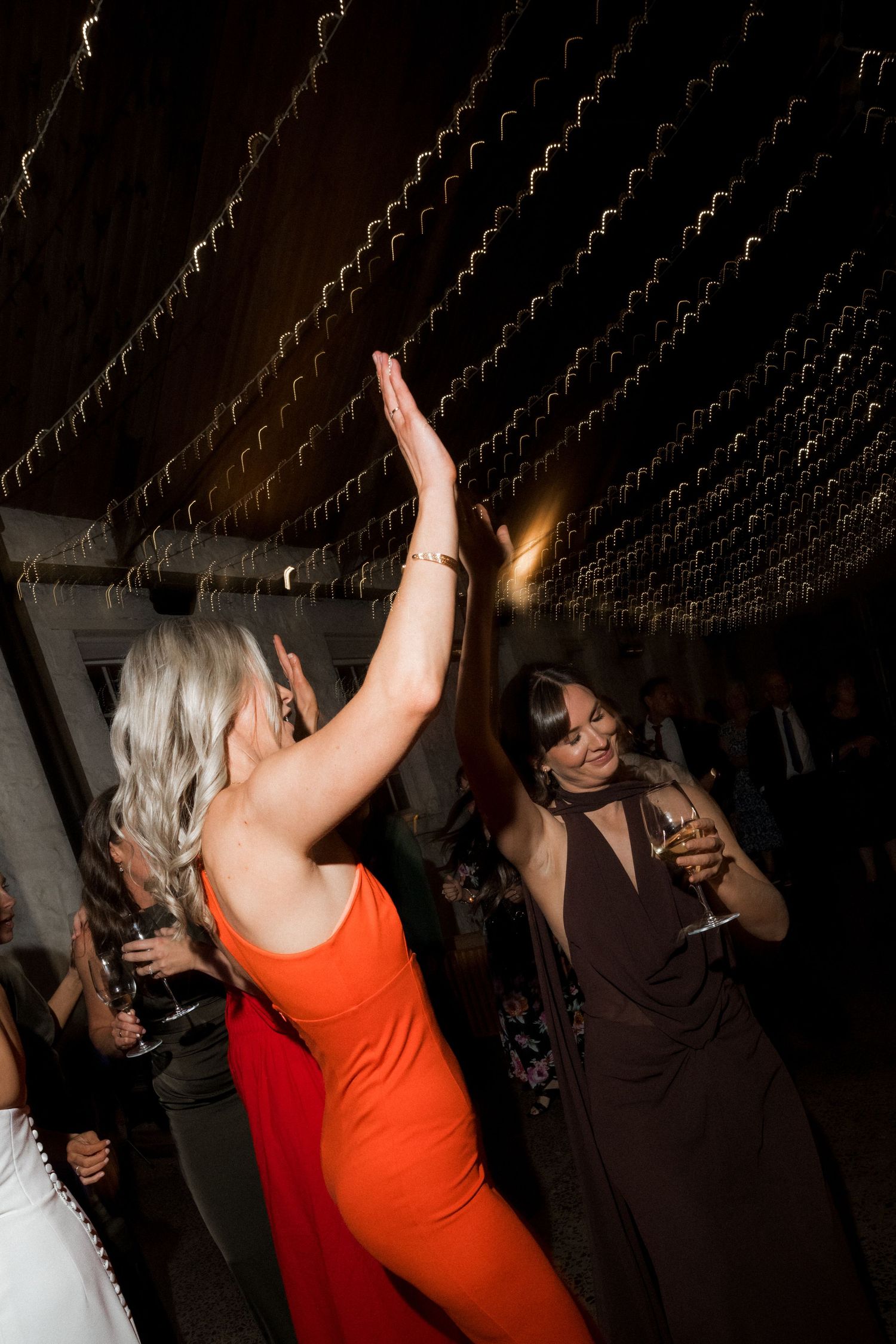 Wedding reception guests dancing and celebrating in dimly lit venue with string lights overhead.