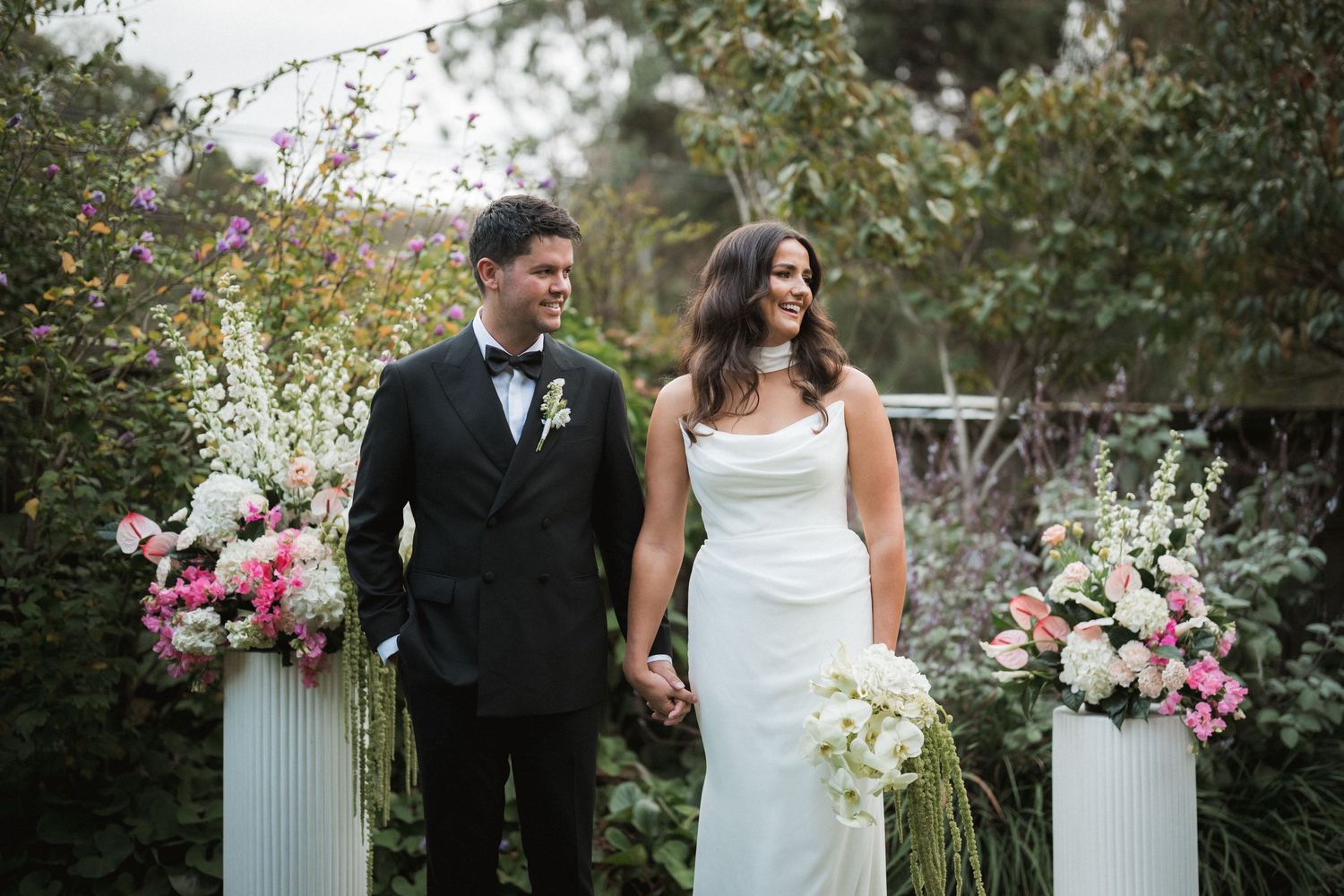 A romantic wedding couple poses together in a lush garden setting surrounded by pink and white floral arrangements.