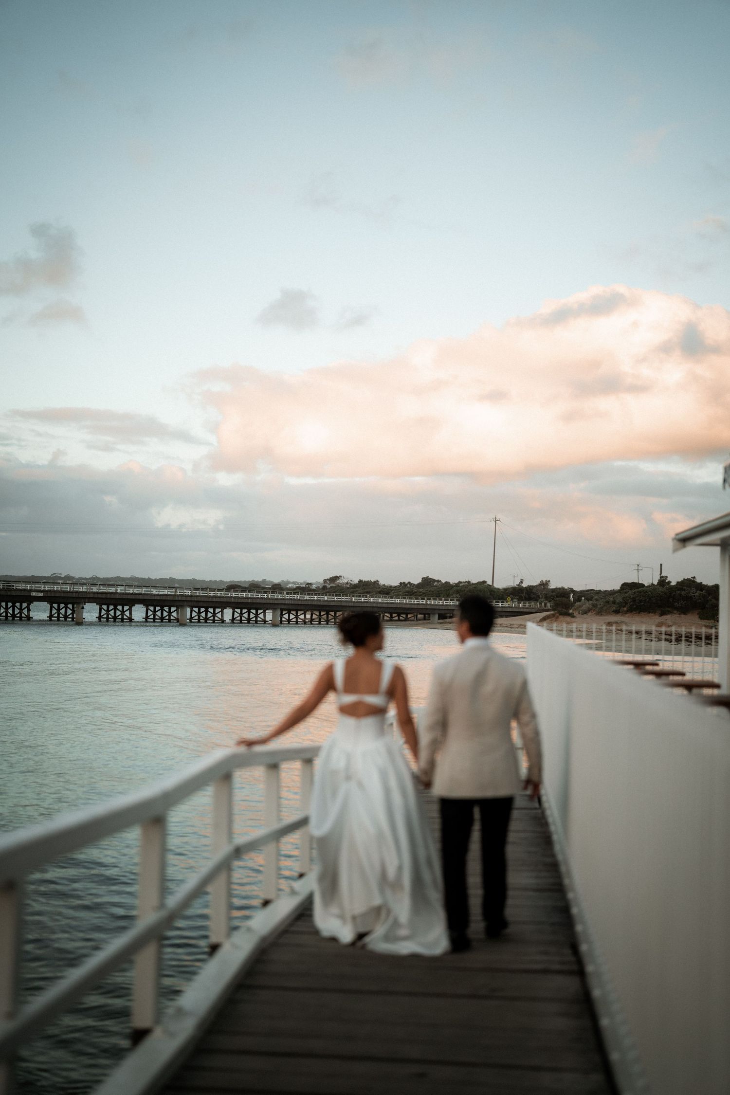 Couple walking along waterfront pier at sunset with ocean and cloudy sky in background.