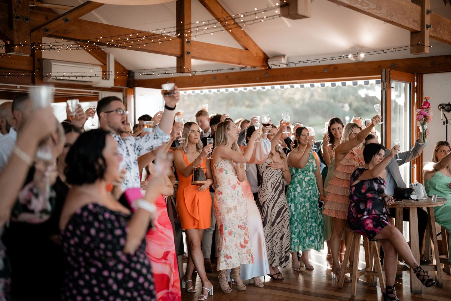 Large group of wedding guests dancing and celebrating in a rustic venue with exposed beams.
