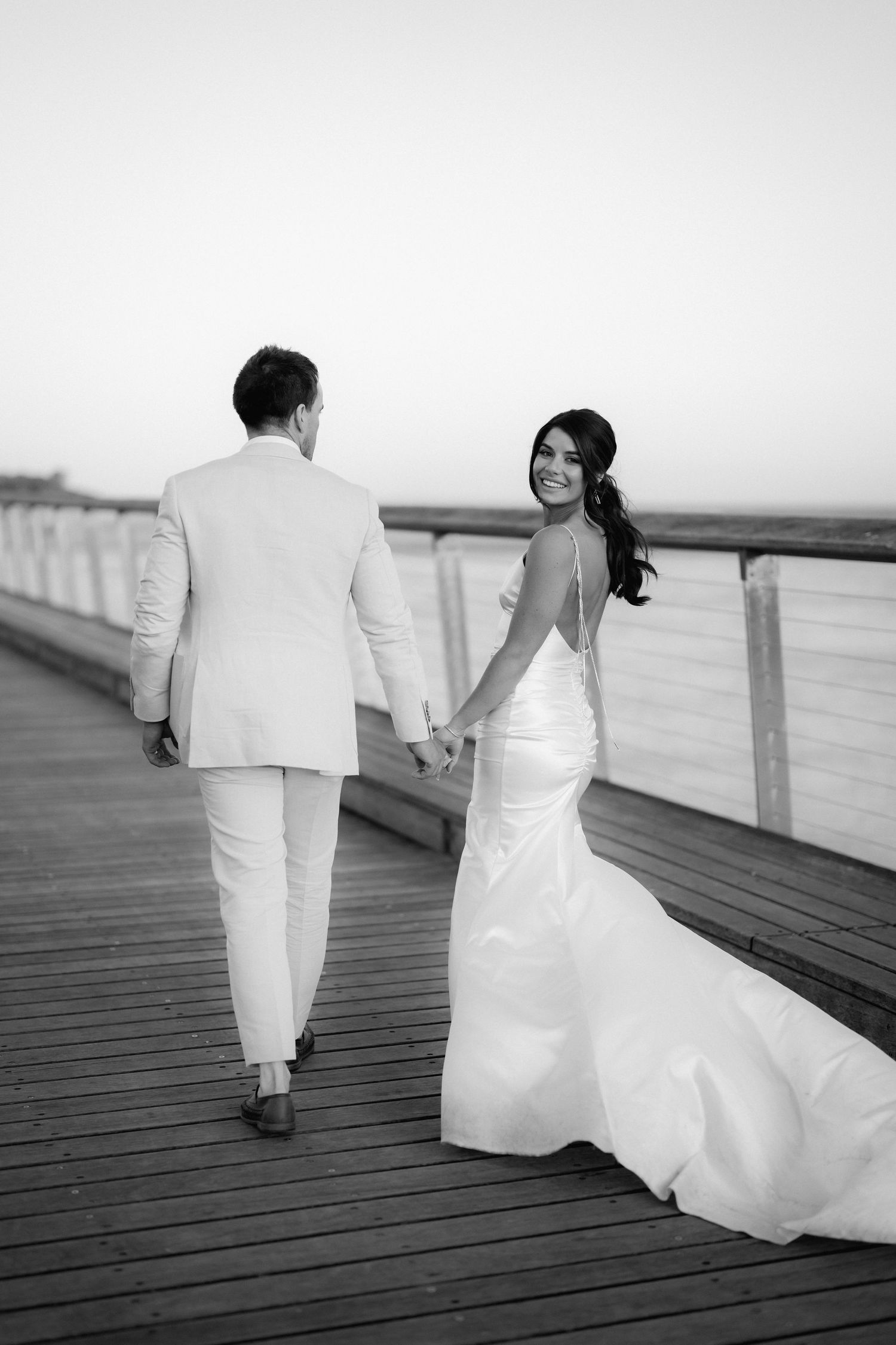 Black and white wedding photos of couple walking on wooden boardwalk by the ocean.