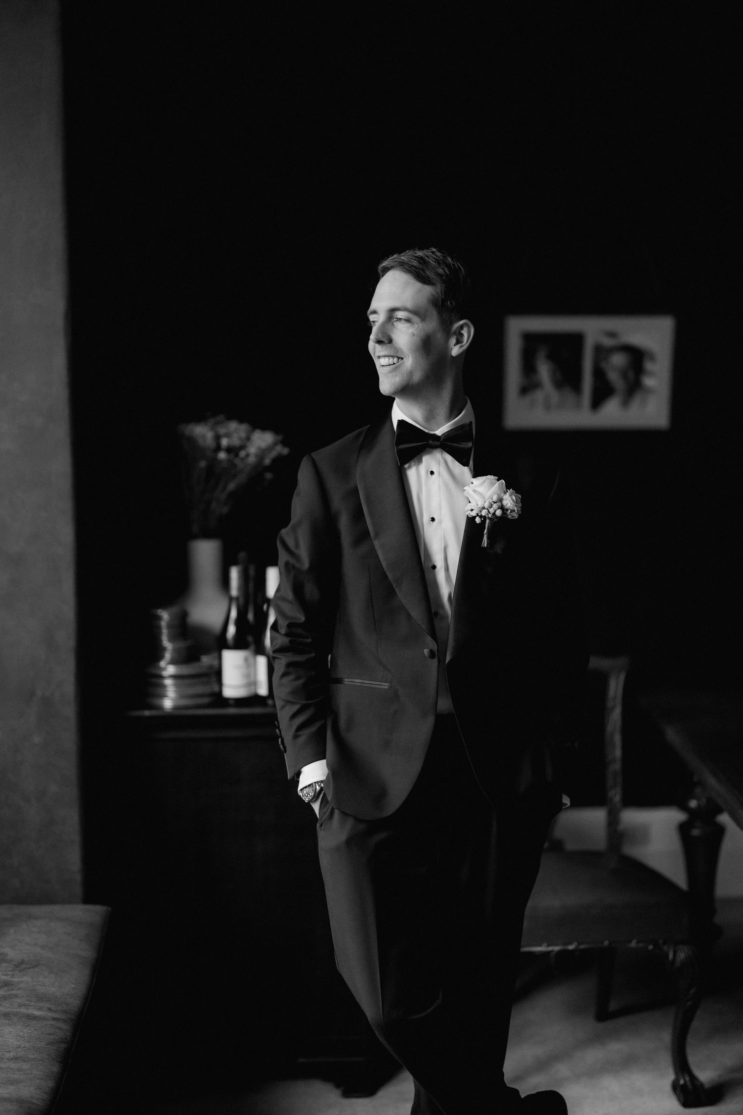 Elegant black and white portrait of a groom in a tuxedo smiling in a dimly lit room.