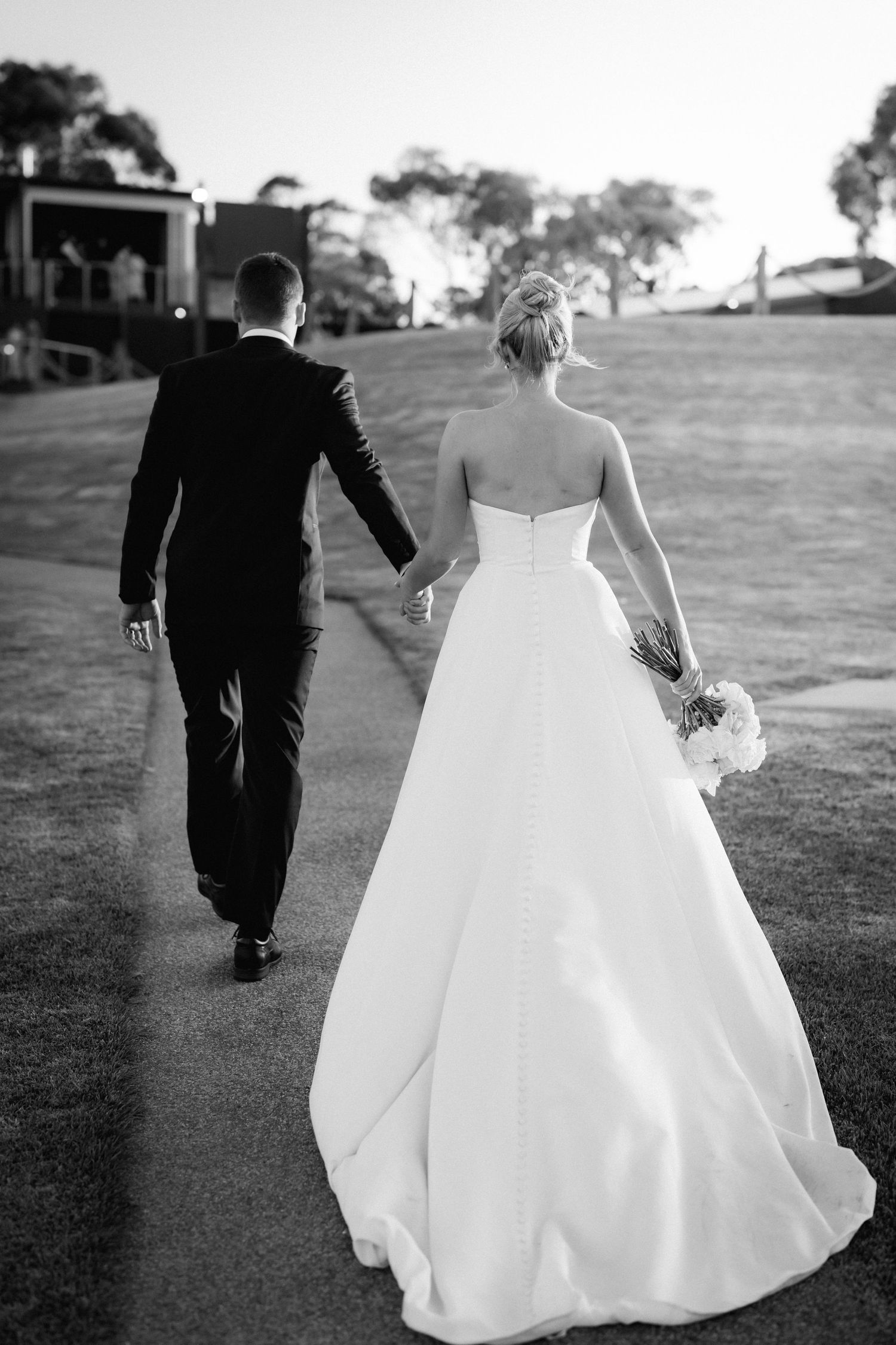 A couple walks hand in hand across a grassy hillside at their wedding, photographed from behind.