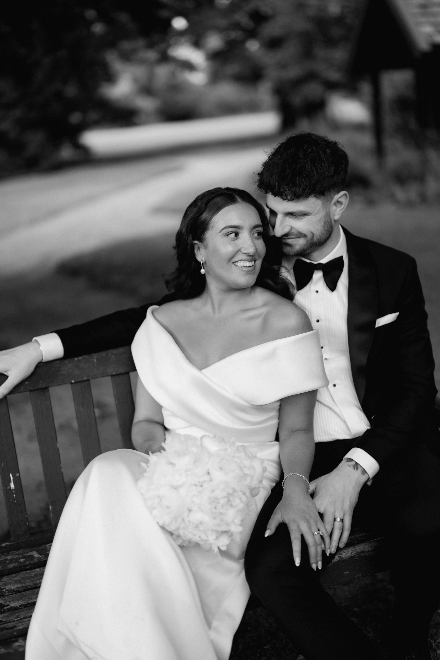 A couple shares an intimate moment while sitting together on a wooden bench in formal attire.