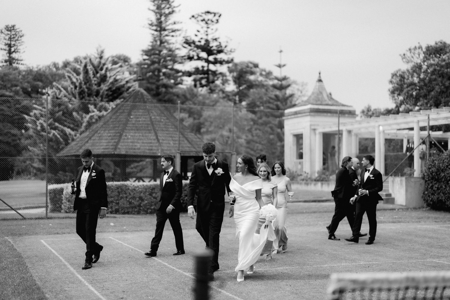 Black and white photo of wedding party walking past a gazebo and tennis courts in a garden setting.