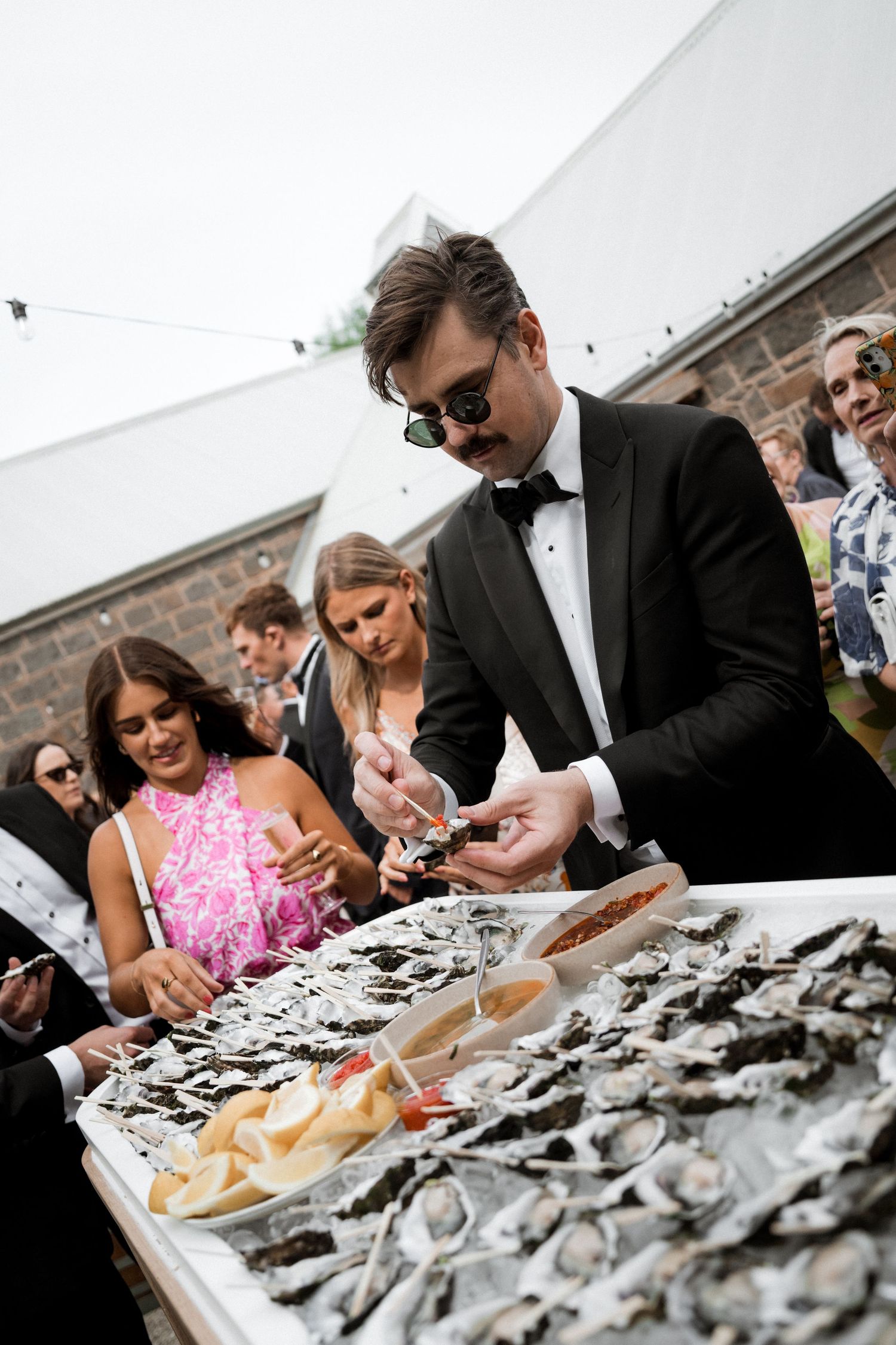 Wedding guests in formal attire gather around a table of fresh oysters at an outdoor reception.