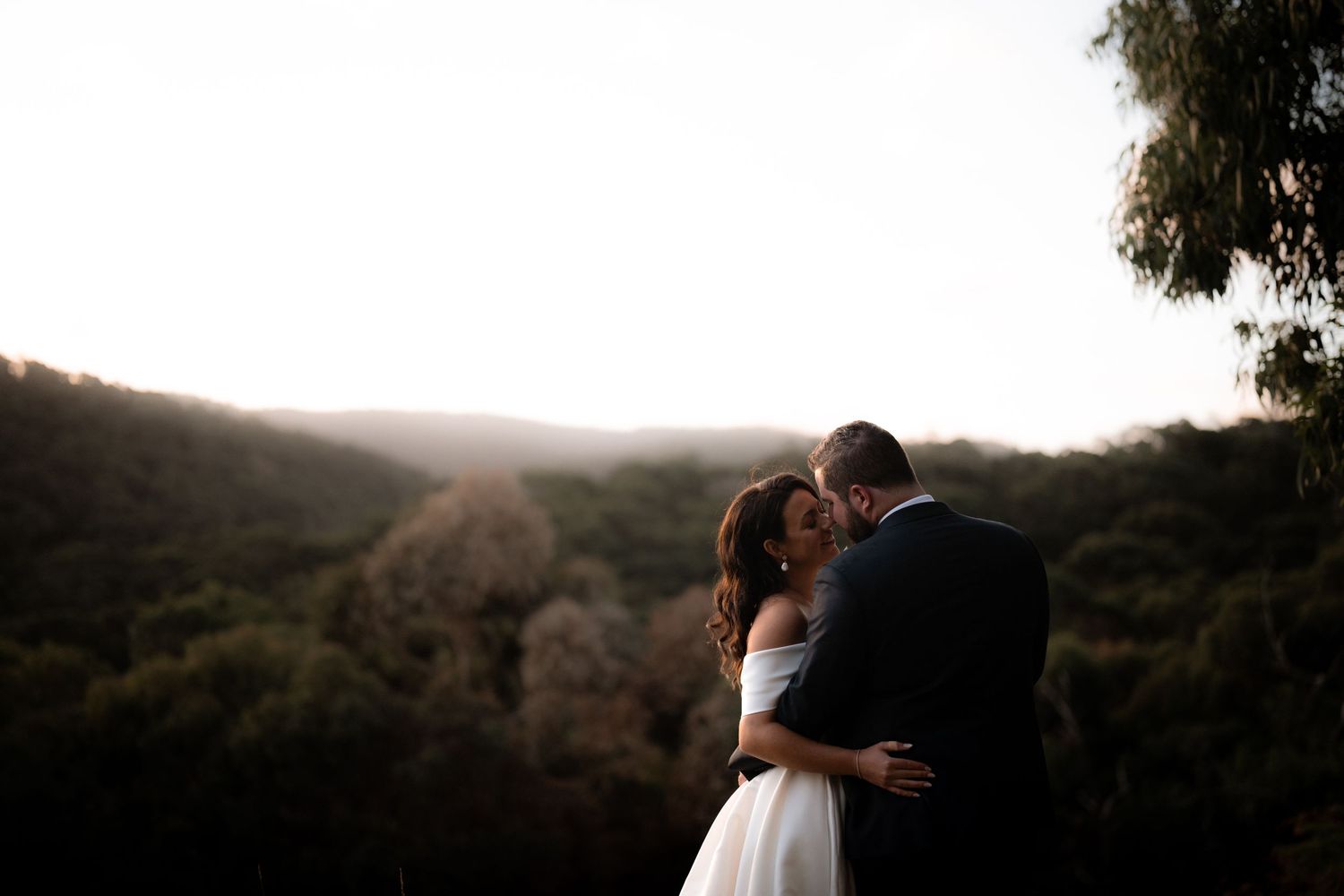 A romantic couple embraces during golden hour on a hillside overlooking a scenic natural landscape.