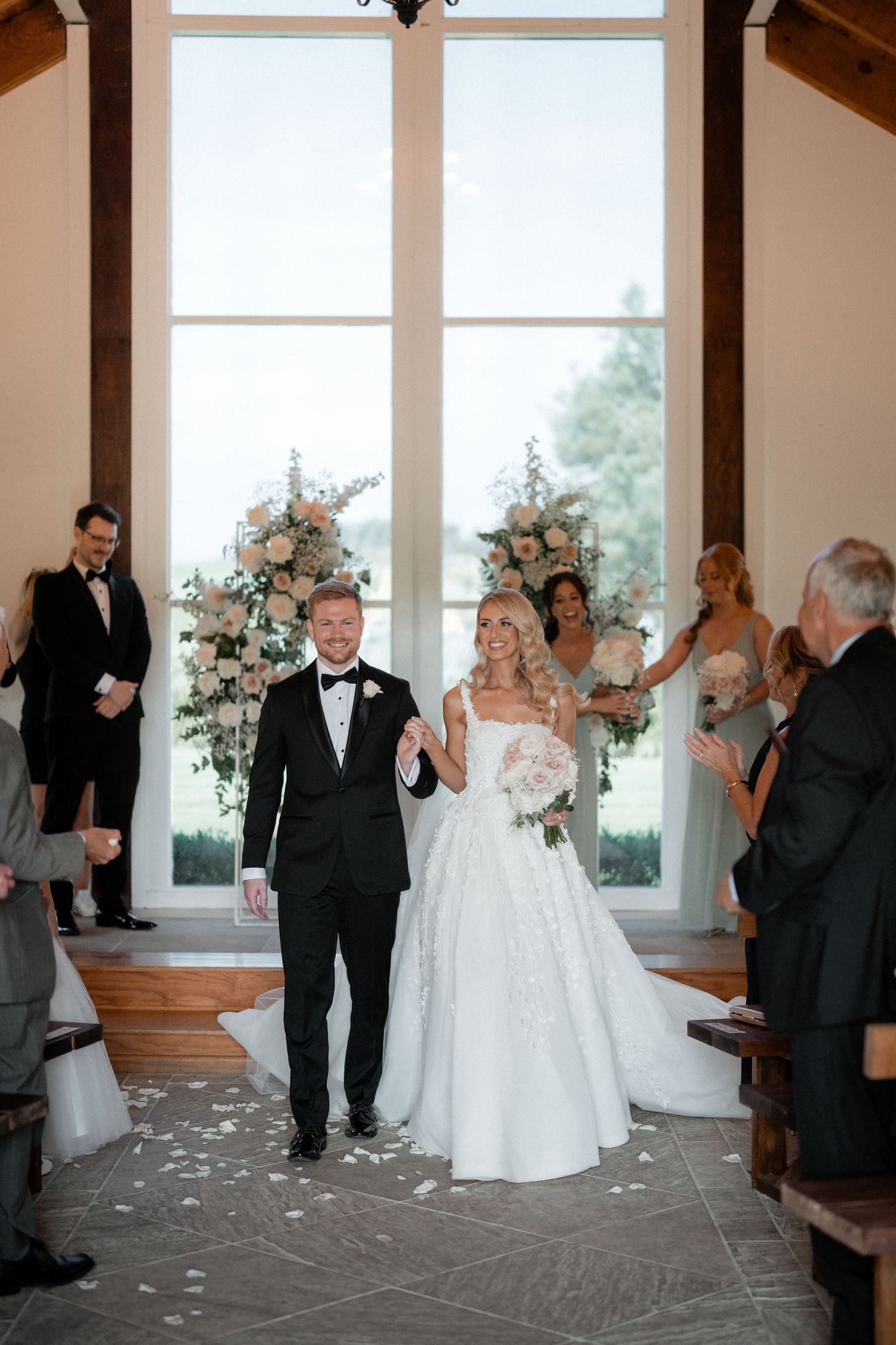 A newlywed couple walking down the aisle of a chapel with floral arrangements and guests.