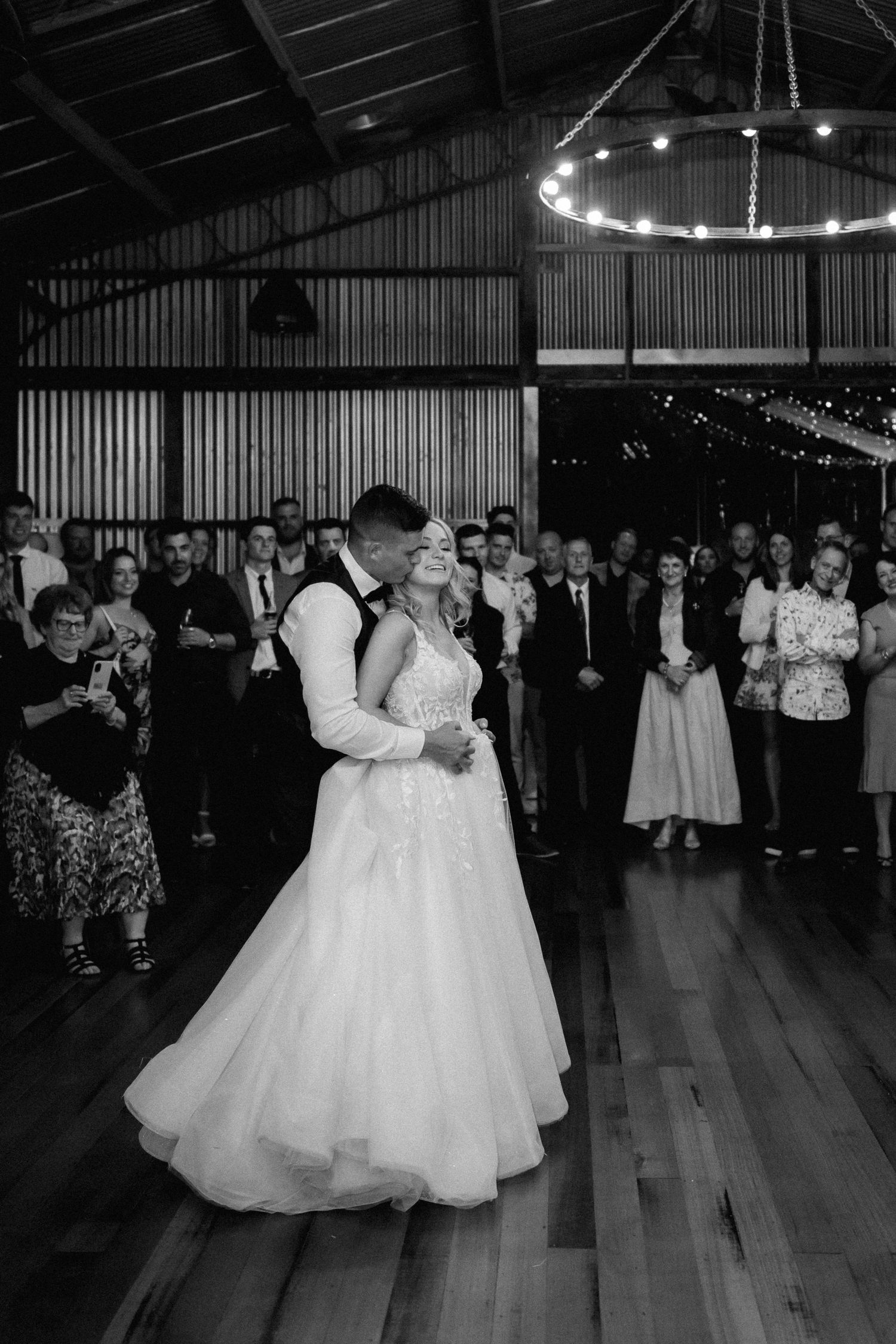 First dance in rustic barn venue with circular chandelier and watching guests.