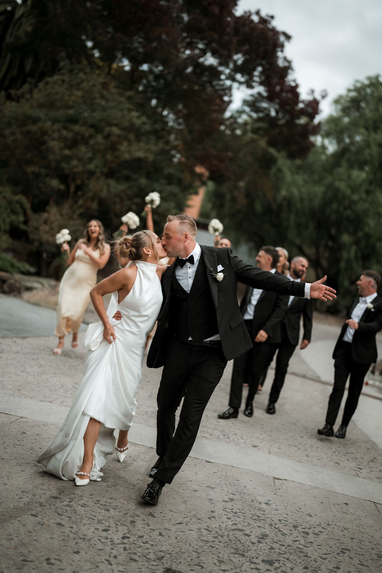 Wedding party strikes playful poses while walking down a tree-lined path during autumn celebrations.