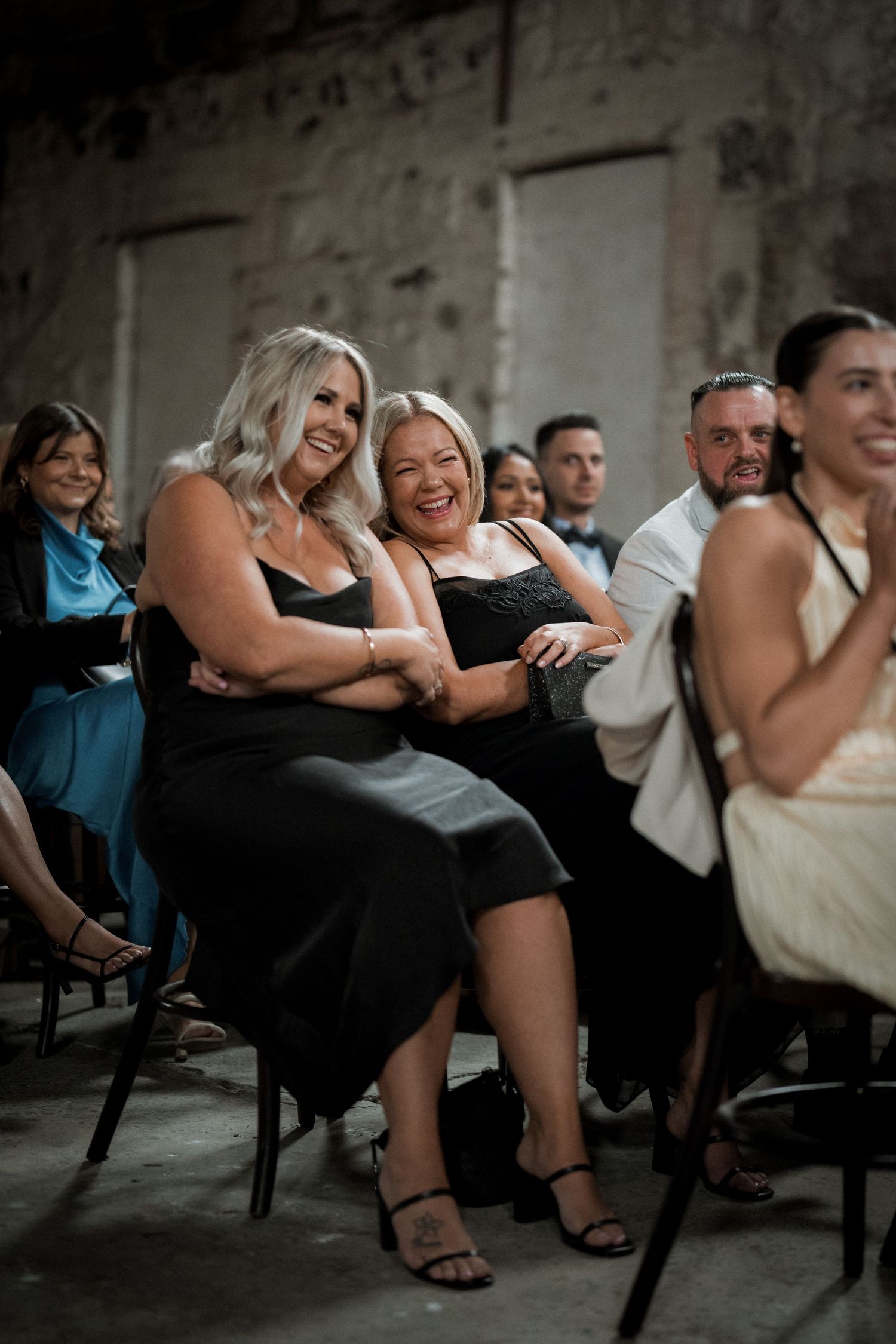 Wedding guests share heartfelt laughter during reception speeches in a rustic venue.