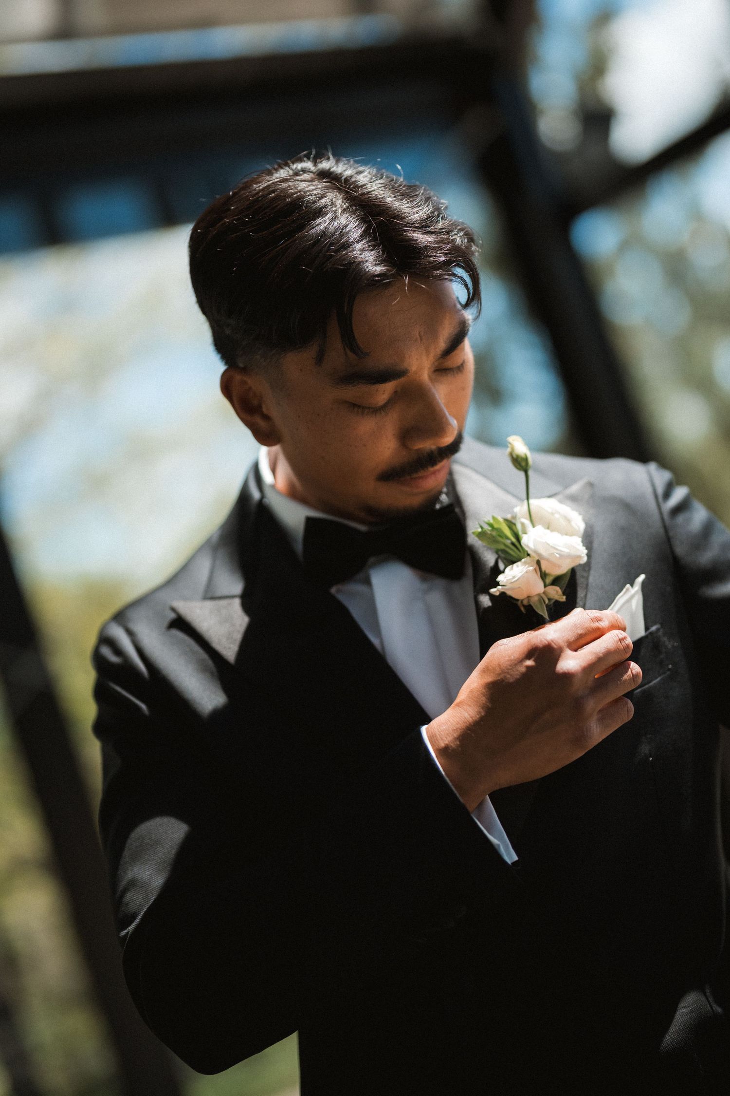 A groom in a black tuxedo and bow tie adjusts a white rose boutonniere on his lapel before the wedding ceremony.