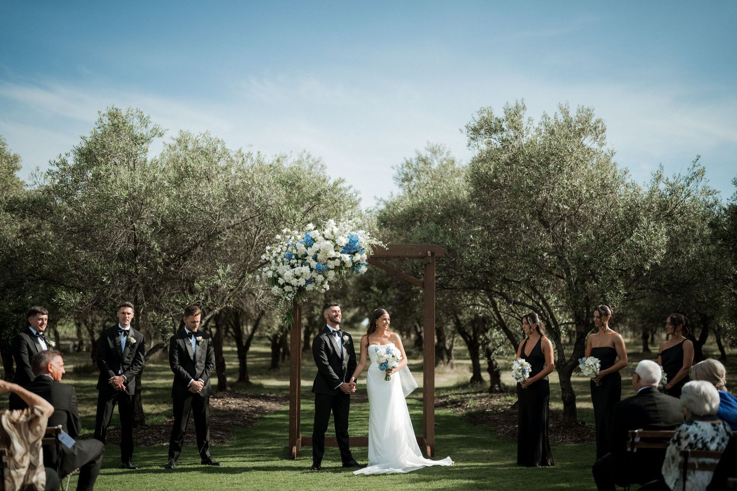 An outdoor wedding ceremony takes place in a scenic olive grove under blue skies on a sunny day.