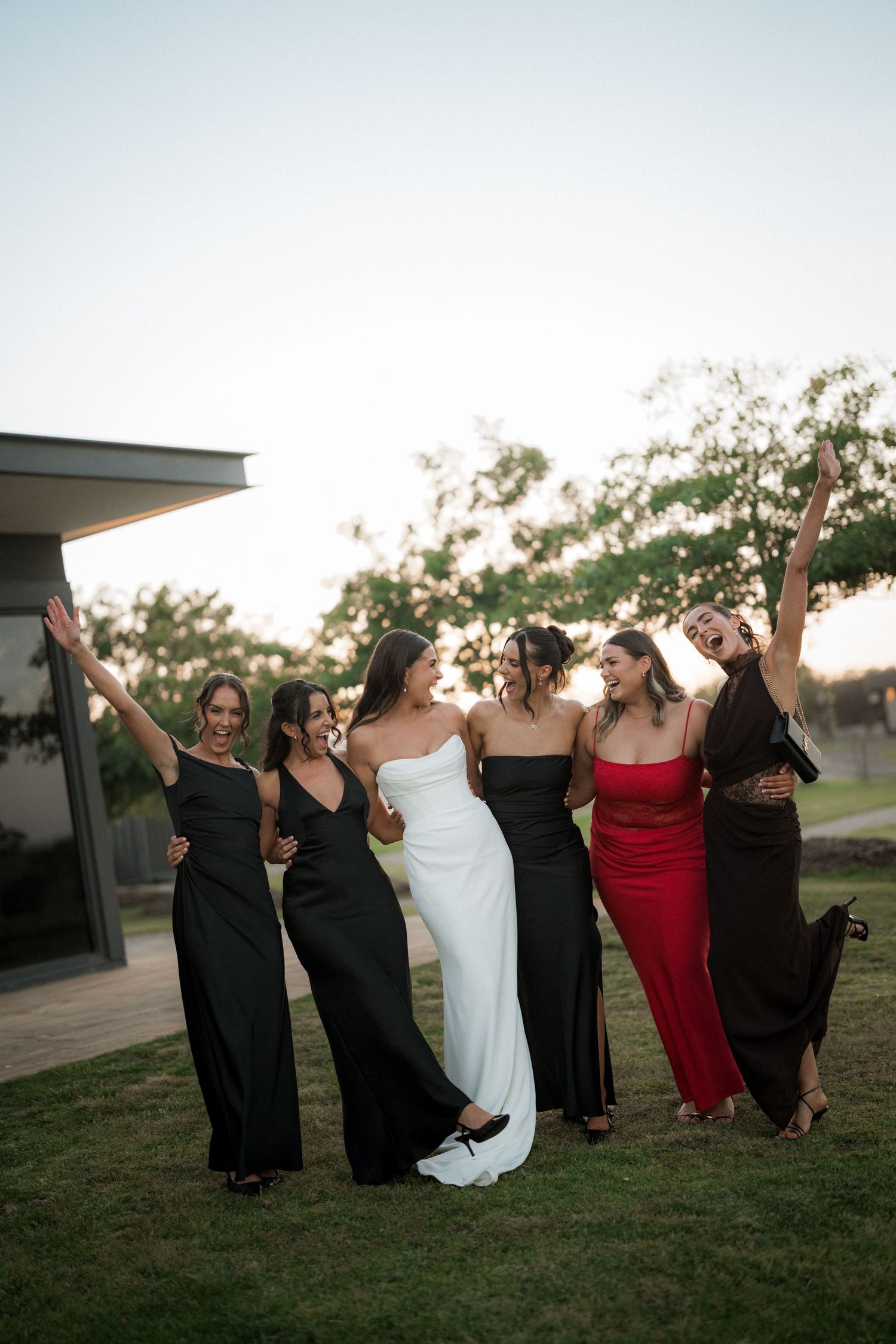 Bride in white gown celebrates with bridesmaids in dark dresses at sunset.