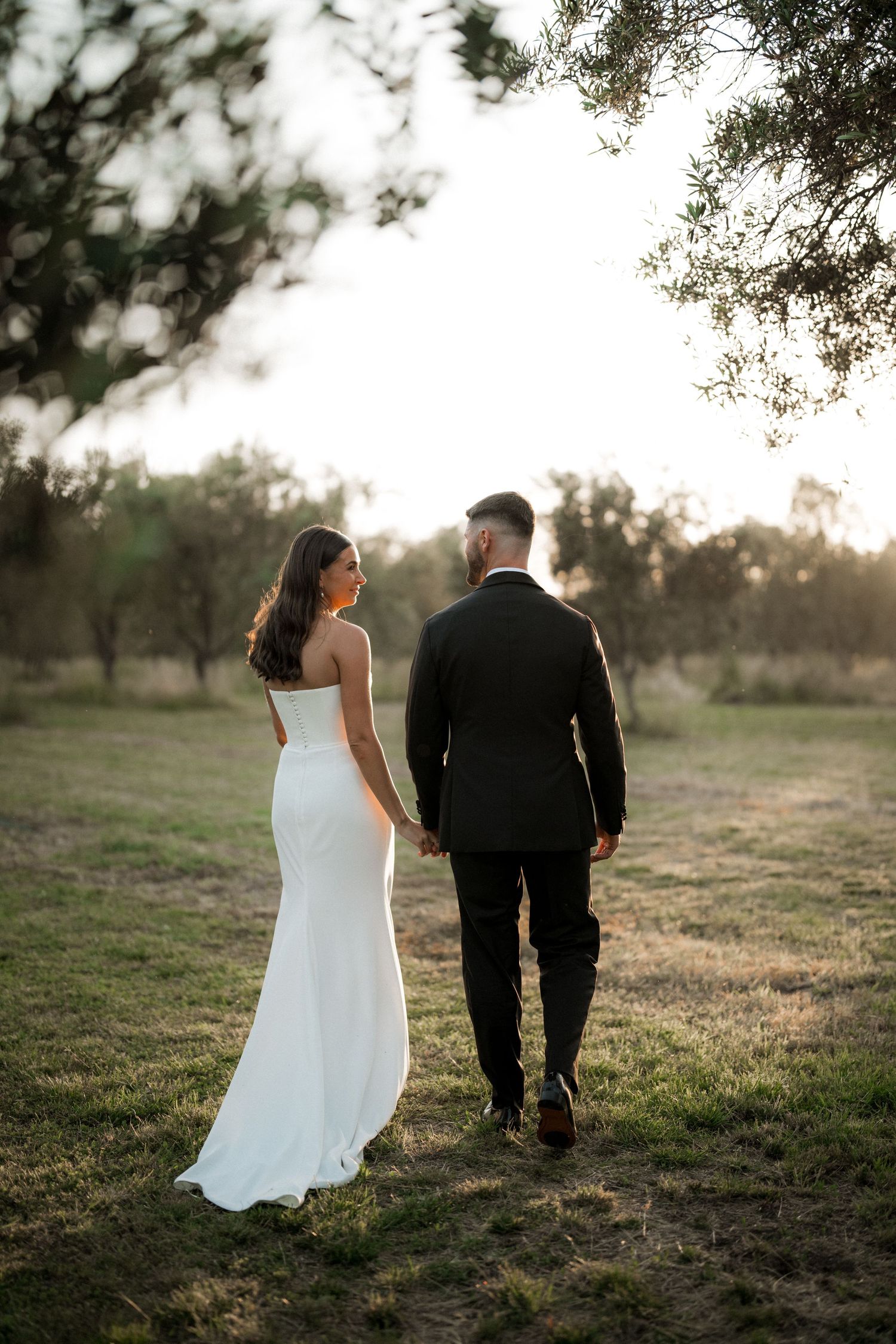 Couple in wedding attire walking through a sunset-lit field.