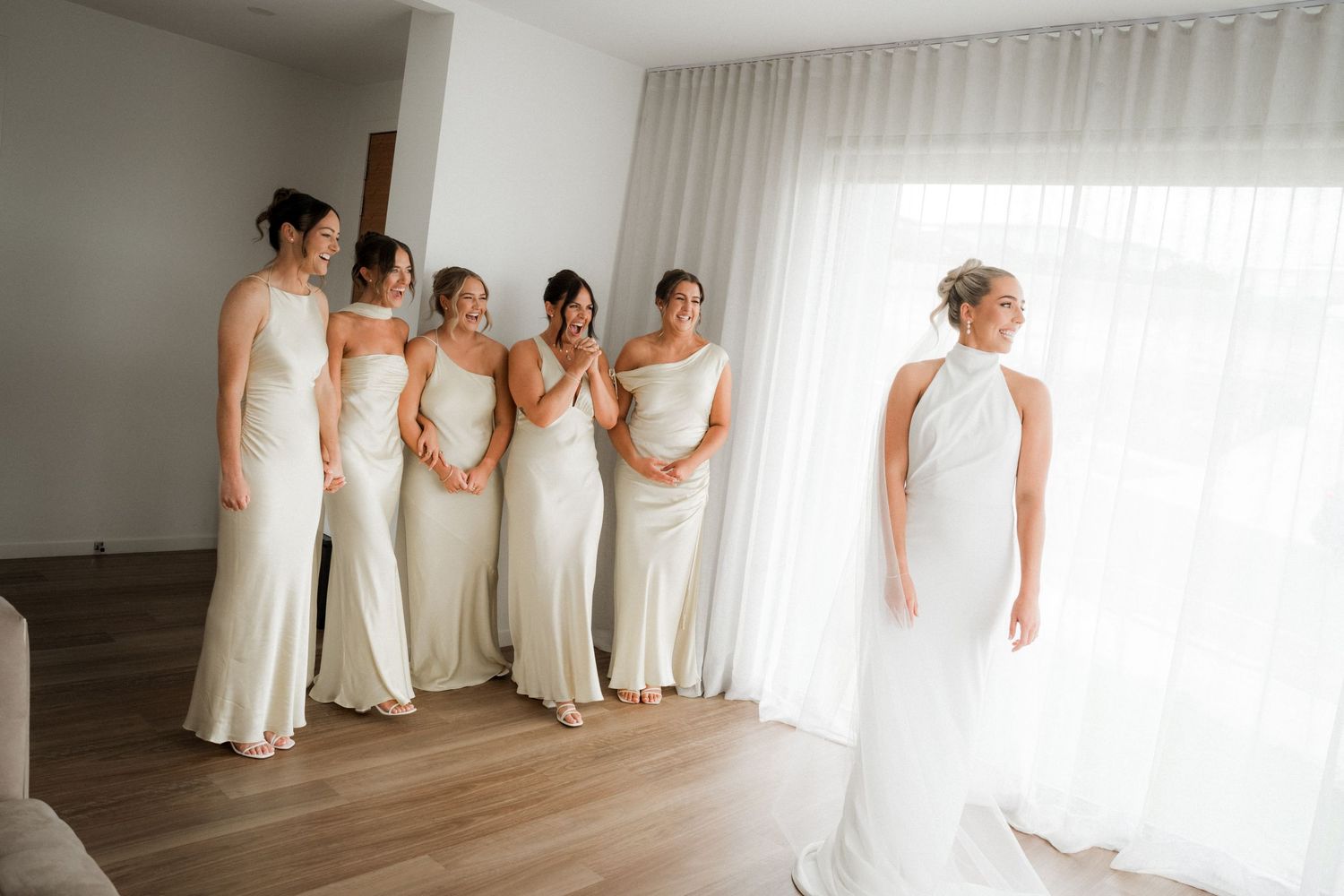 Bridesmaids in flowing white dresses pose together near a window in a bridal suite.