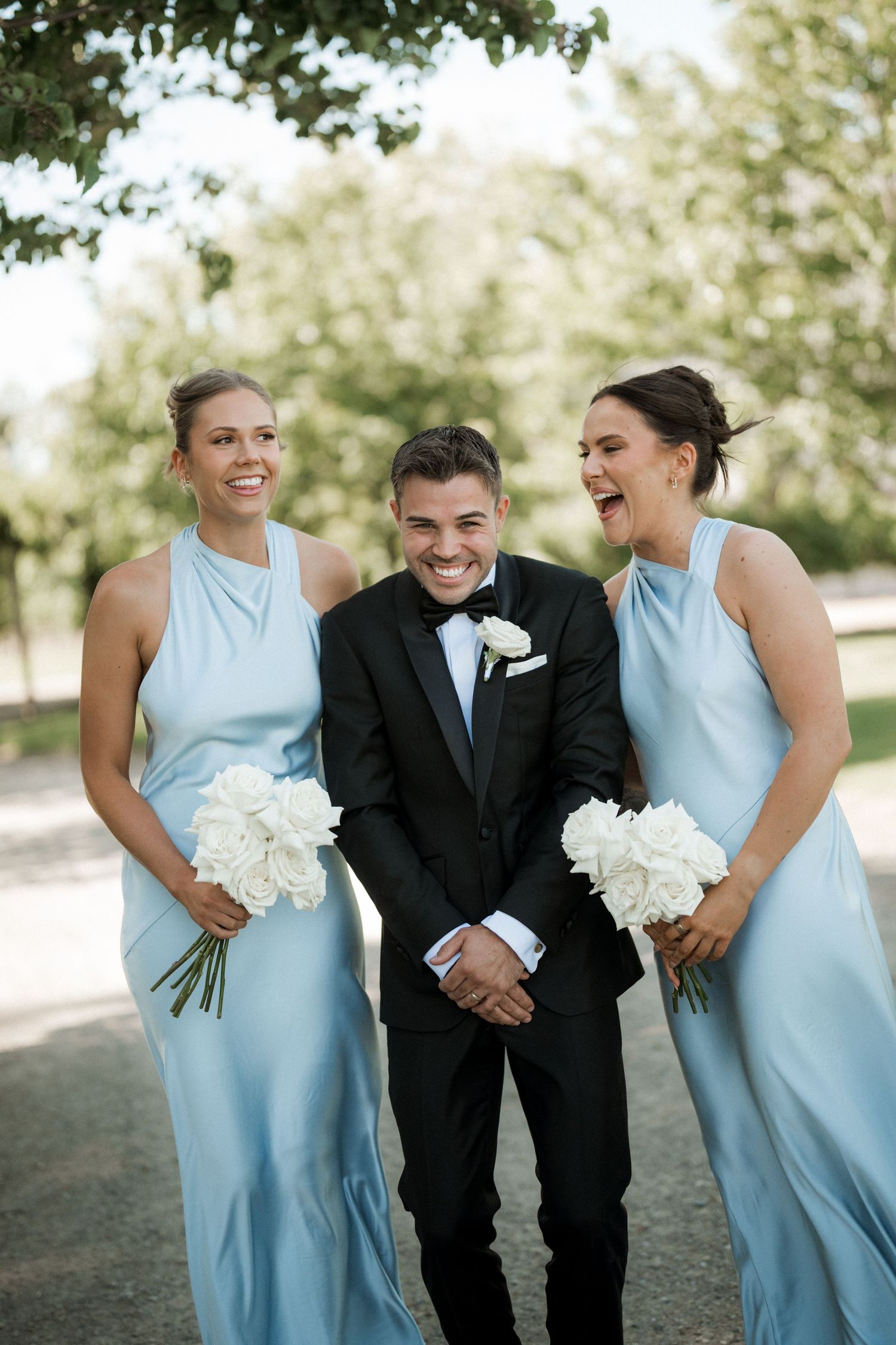 Wedding party in light blue dresses and black tuxedo sharing laughs outdoors.