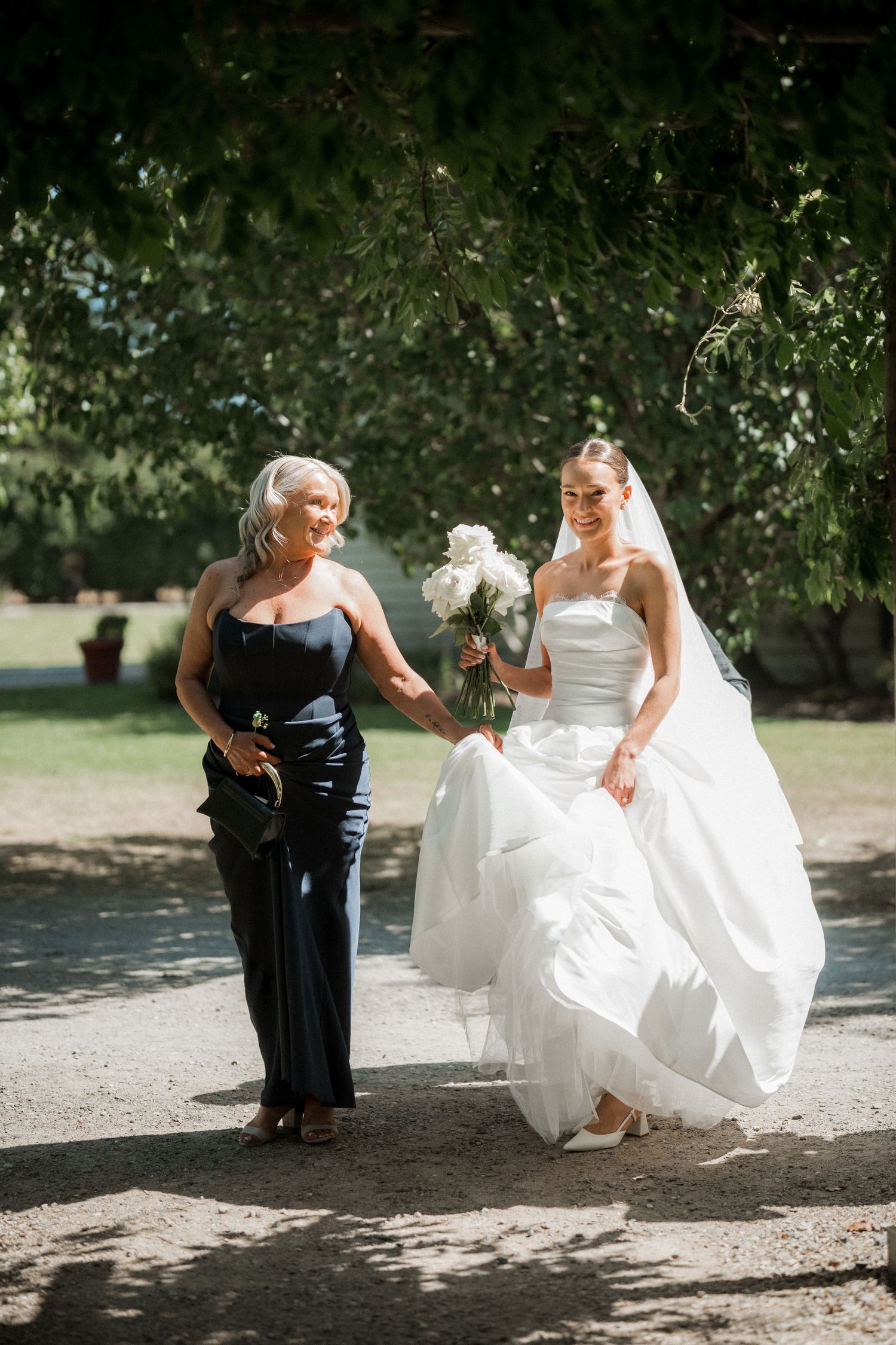 Elegant wedding scene with flowing white dress and black gown under dappled sunlight in garden setting.