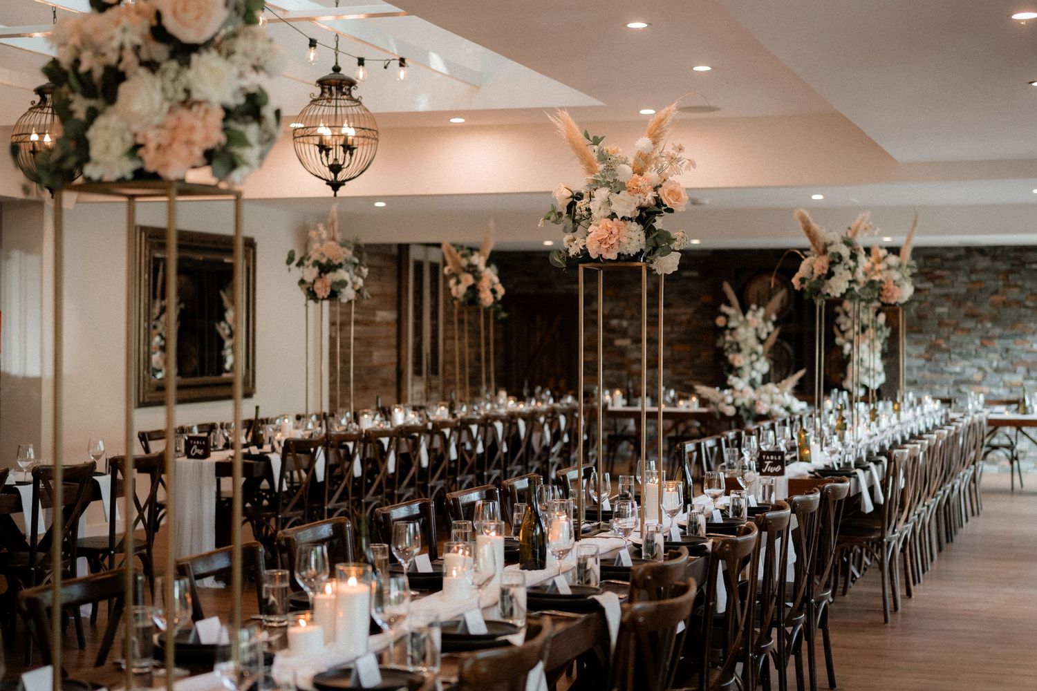 Long banquet tables with elegant pink floral centerpieces and chandeliers are arranged in a rustic wedding reception venue.
