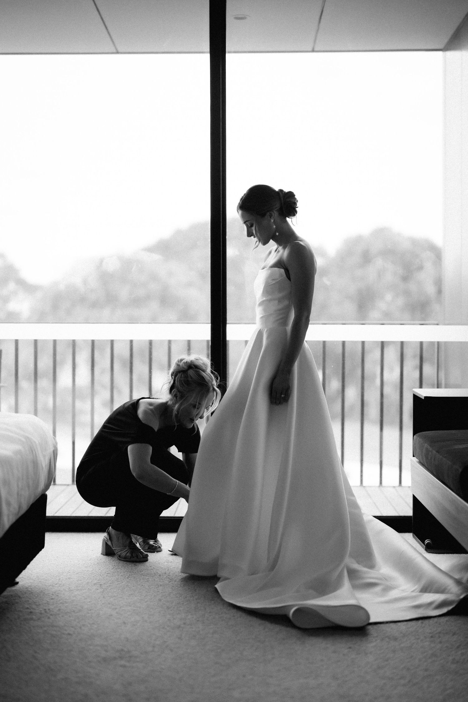 Black and white photo of final bridal preparations with a person adjusting the hem of an elegant white wedding gown.