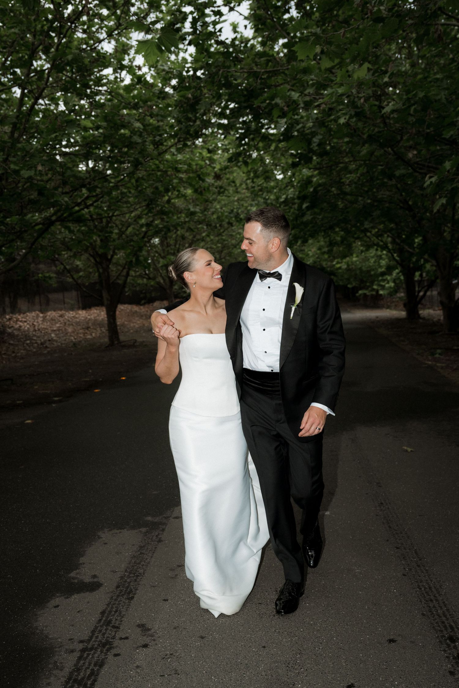 A bride and groom walk hand in hand through a tree-lined path on their wedding day.