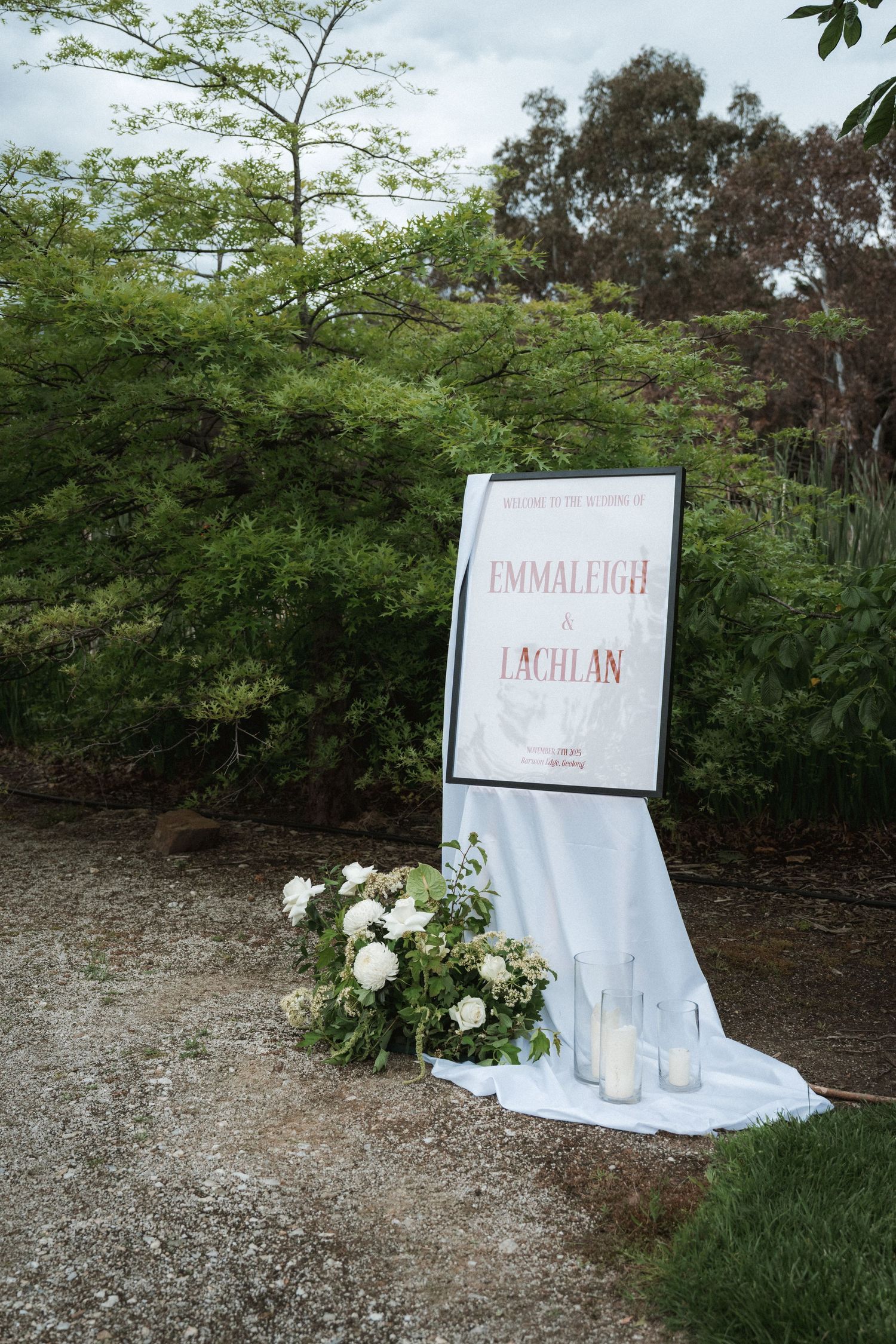Elegant wedding welcome sign for Emmaleigh and Lachlan, adorned with white florals and candles against lush greenery.