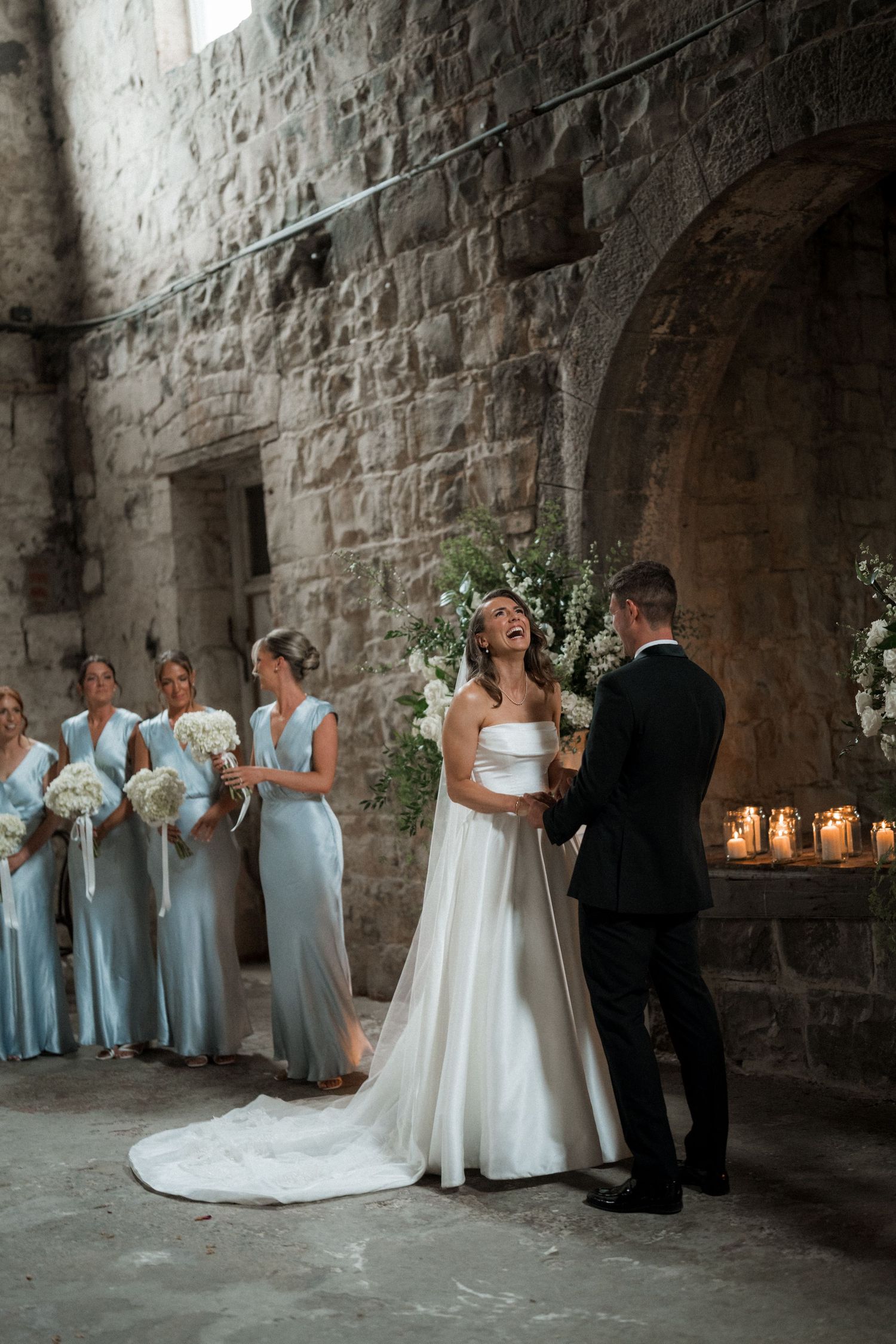 A wedding ceremony in a stone castle with bridesmaids in light blue dresses watching the couple exchange vows.