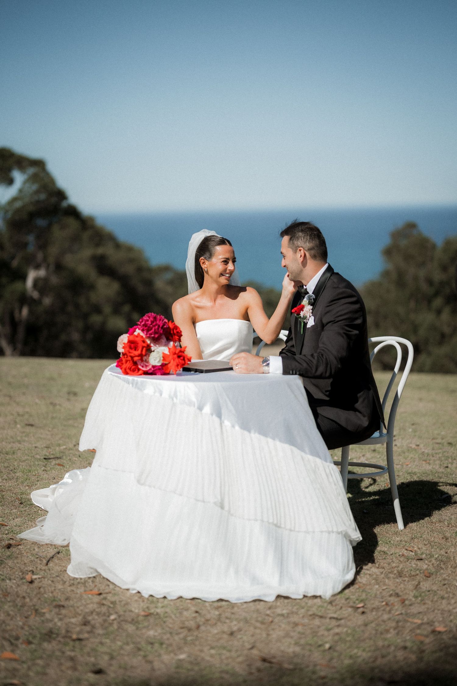 Newlyweds sit at an outdoor table with red floral decorations overlooking the ocean.
