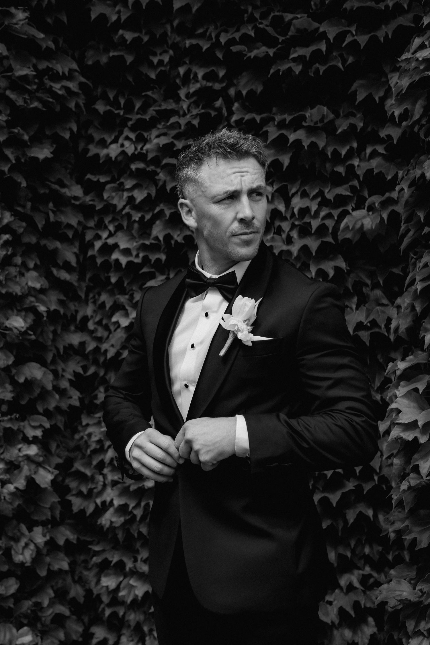 Close-up detail shots of groom adjusting tuxedo cuff and boutonniere against ivy background.