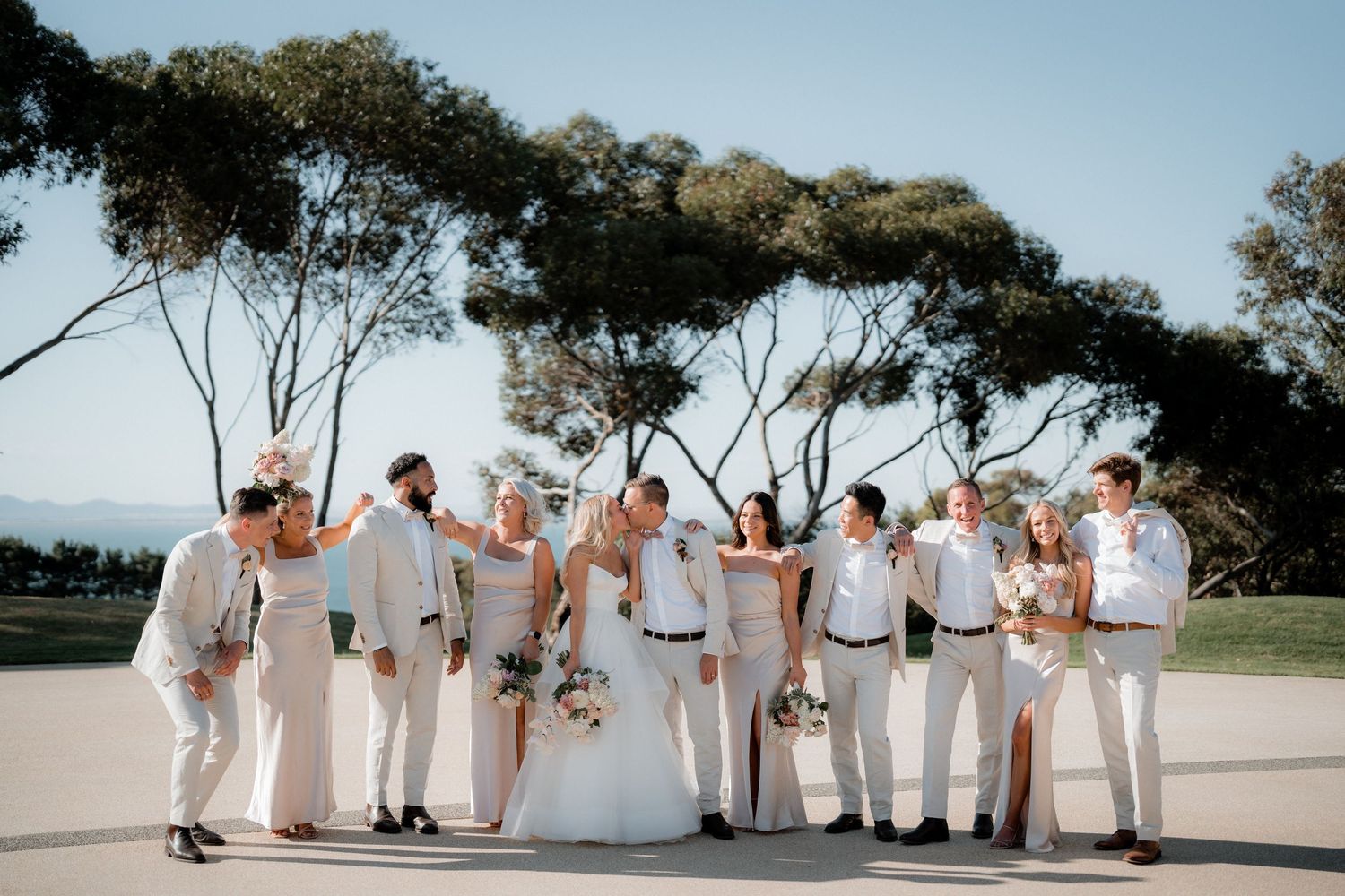 Wedding party lined up for formal photos outdoors under blue sky and trees.