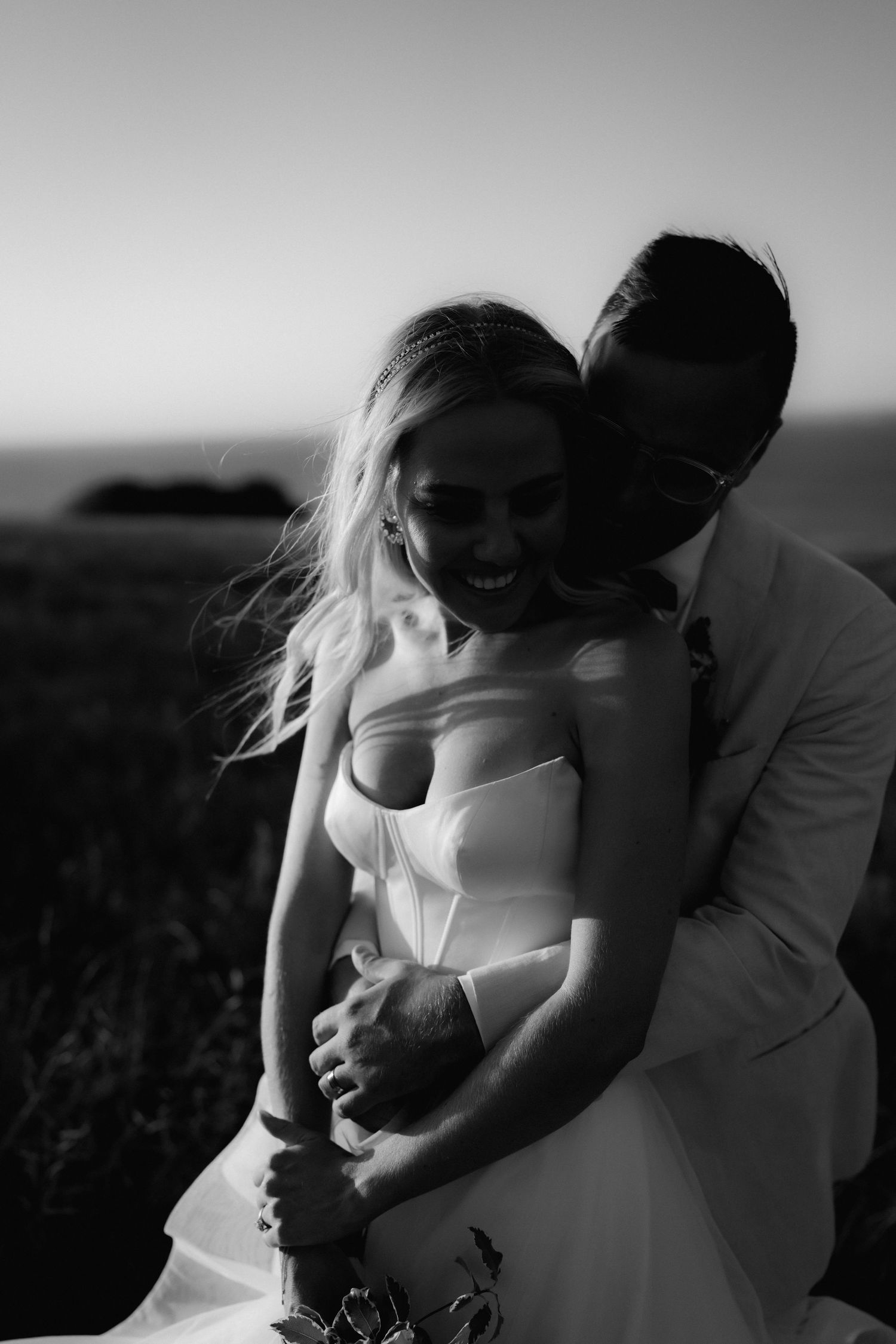 Romantic black and white portrait of a couple embracing in a field at sunset.