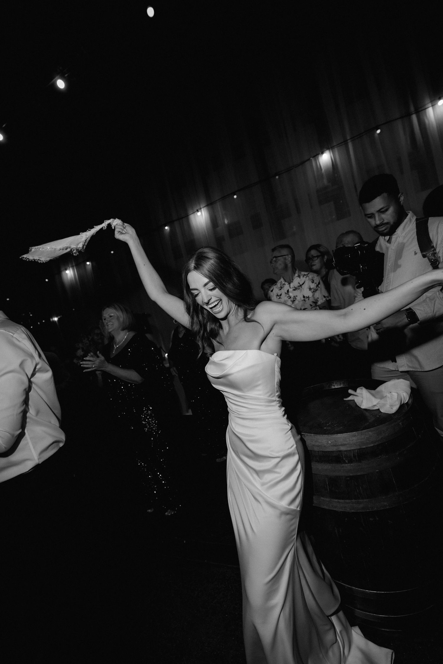 A bride in a white dress dances joyfully at a wedding reception in a dimly lit venue with guests in the background.