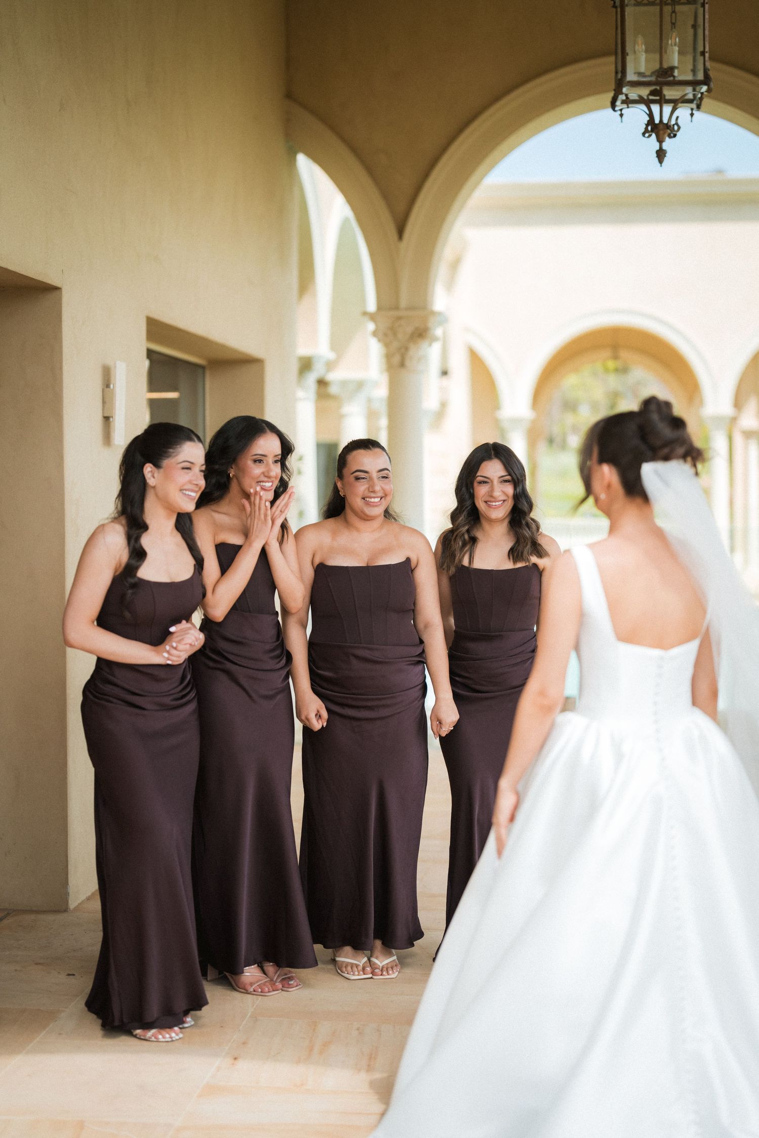 Four bridesmaids in brown satin dresses smile and react joyfully as the bride in a white gown stands before them at a wedding venue.