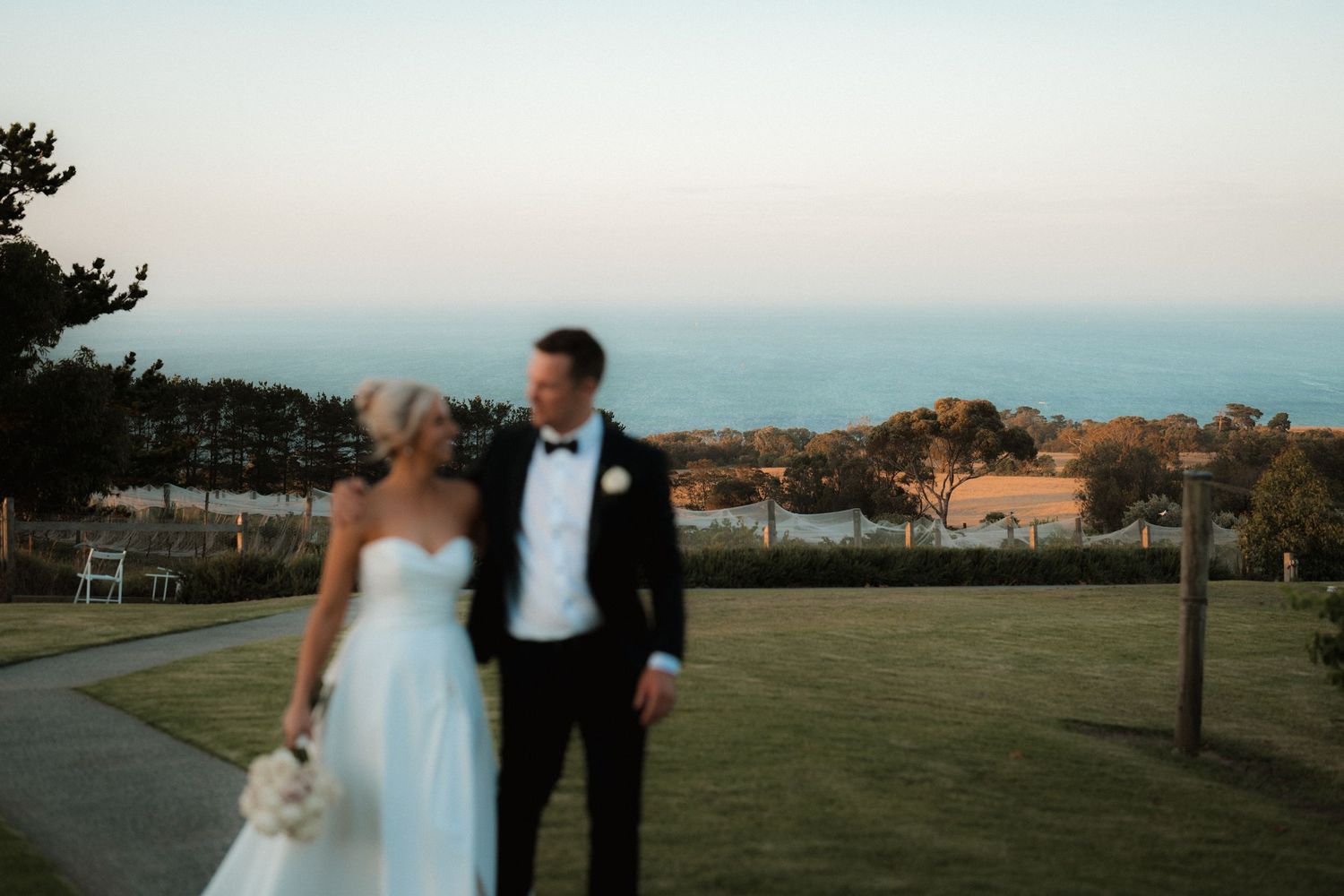 Newlyweds walk together on path with ocean view and sunset sky in background.