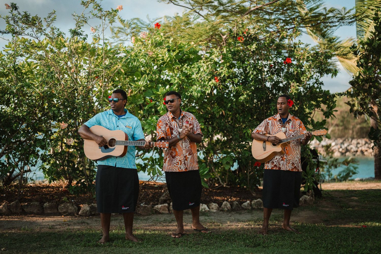 Three musicians in Hawaiian shirts perform with guitars outdoors near tropical foliage.