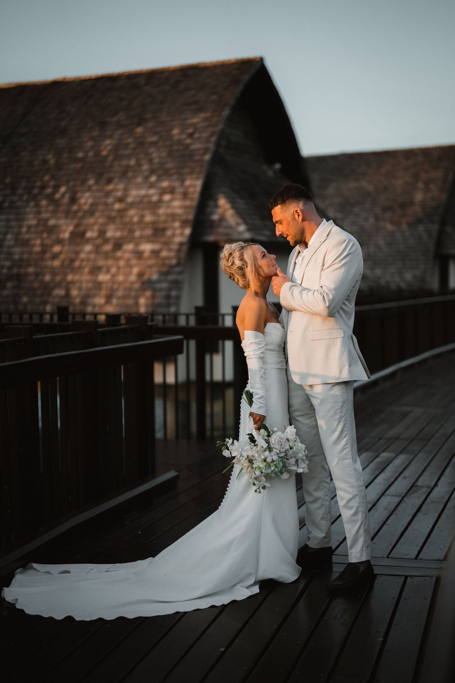 A newlywed couple shares an intimate moment on a wooden deck at dusk with rustic barn architecture in the background.