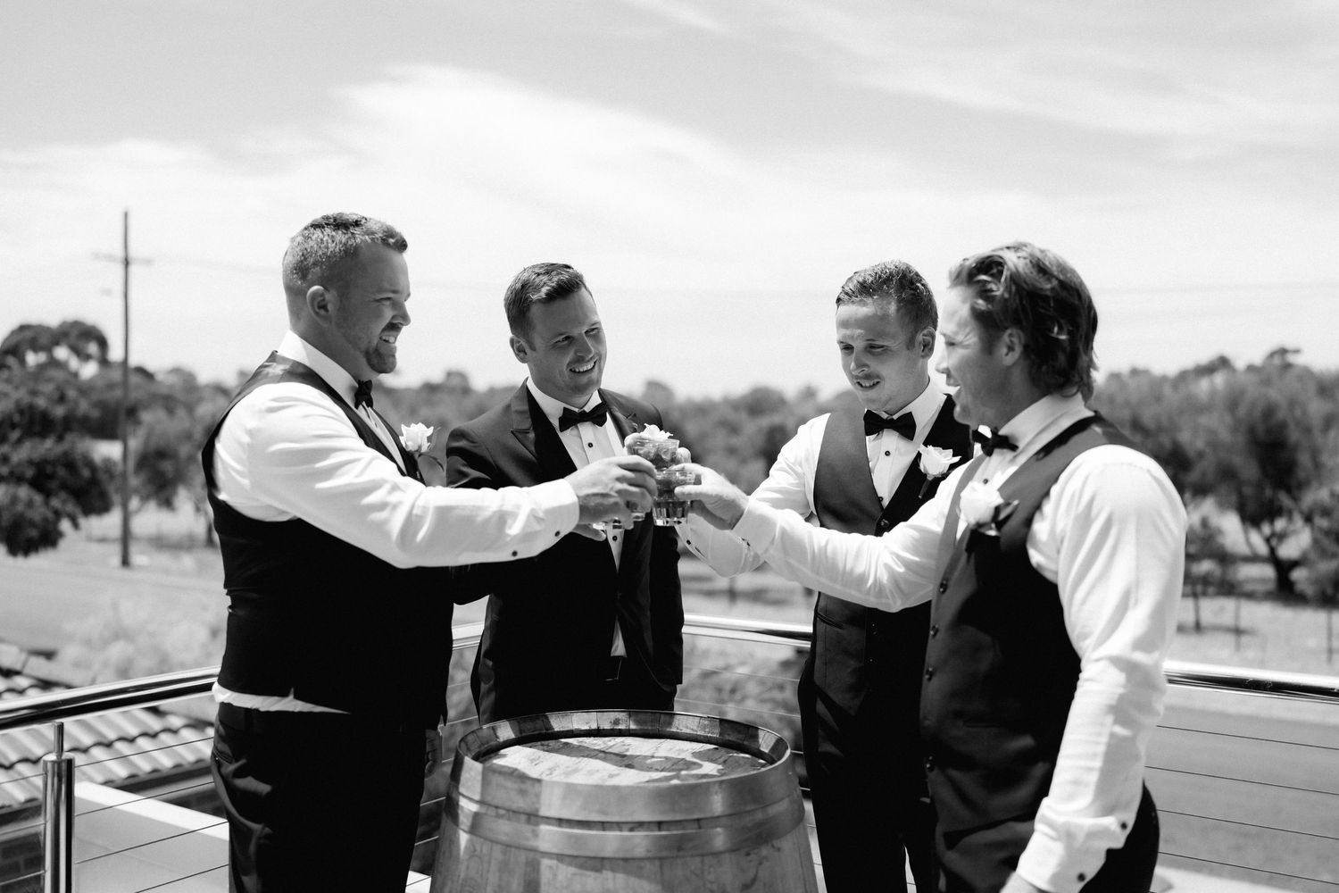 A group of men in vests pour drinks and gather around a barrel in a black and white vintage photograph.