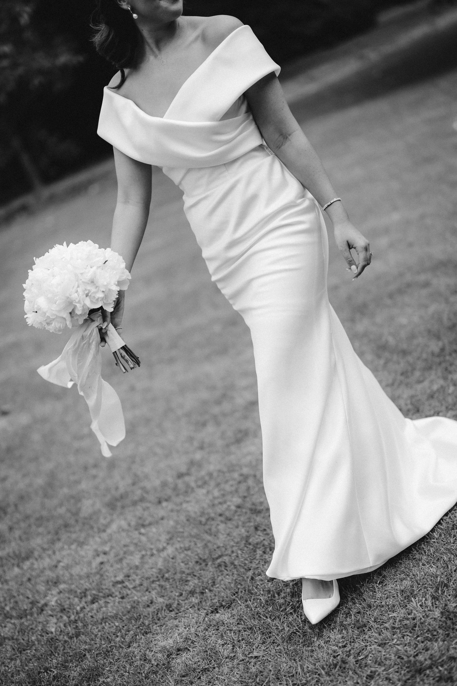 Elegant bride in form-fitting off-shoulder wedding gown holds white bouquet while walking on grass in black and white photo.