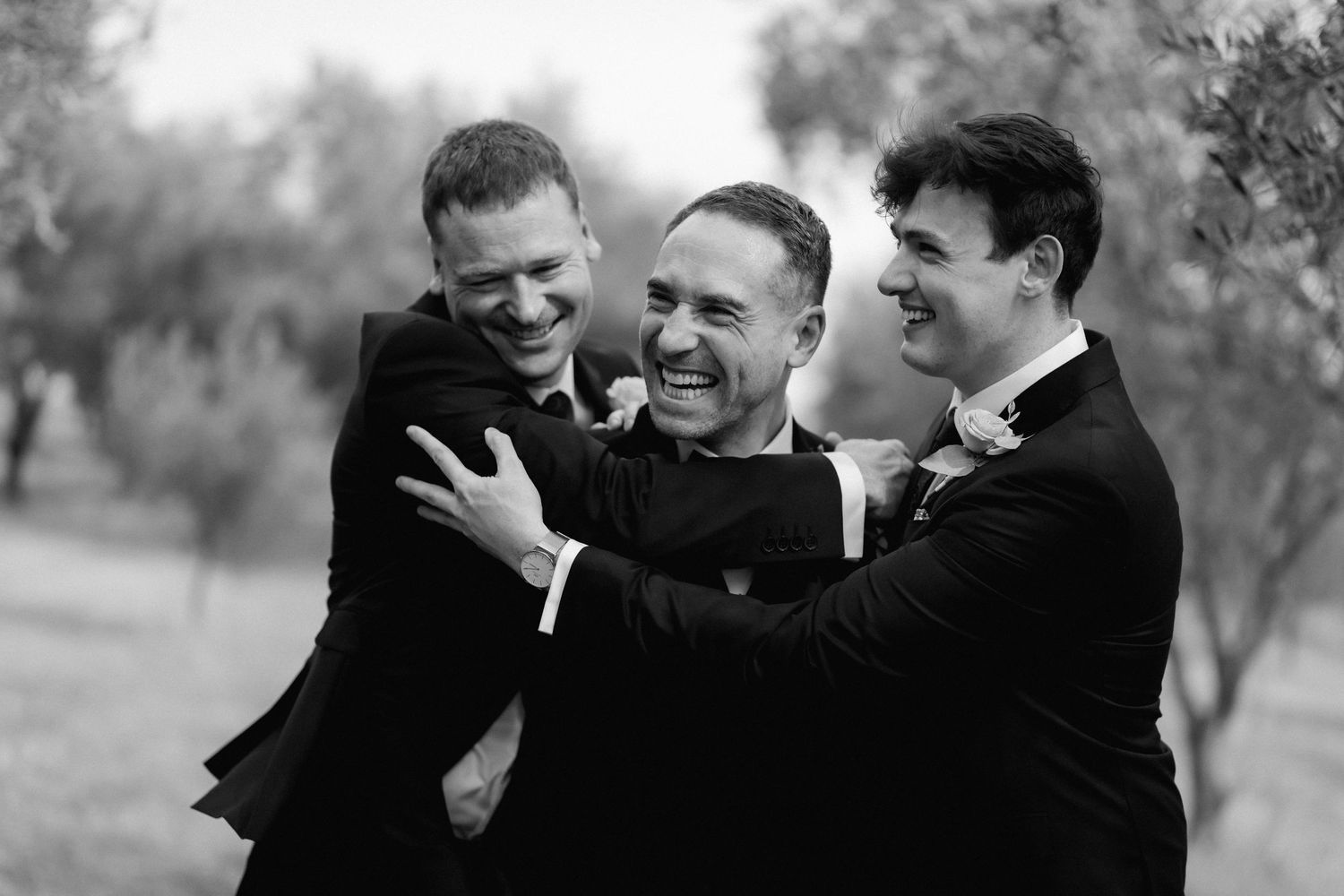 Three people in black suits share a joyful candid moment laughing together outdoors in a natural setting.